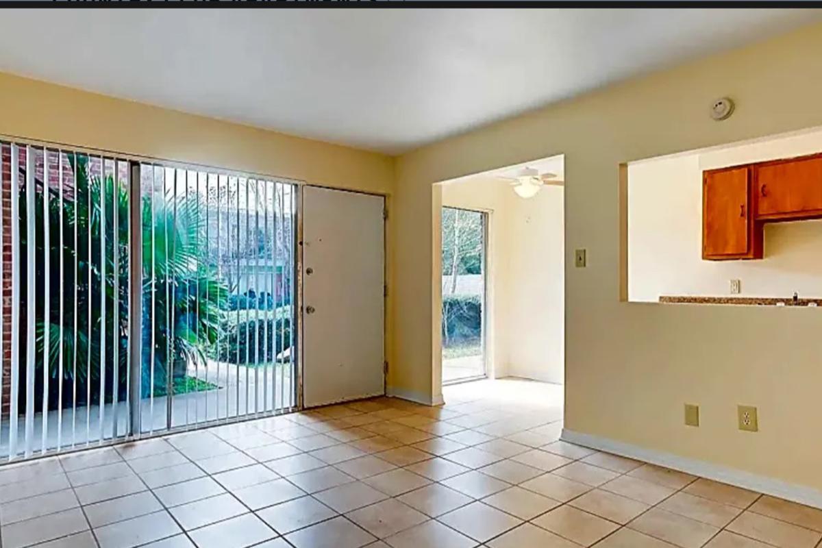 A bright, empty room with tiled flooring, featuring large sliding glass doors that lead to an outdoor area with greenery. To the right, there's an opening to a kitchen area with wood cabinets. Natural light illuminates the space, creating a welcoming atmosphere.