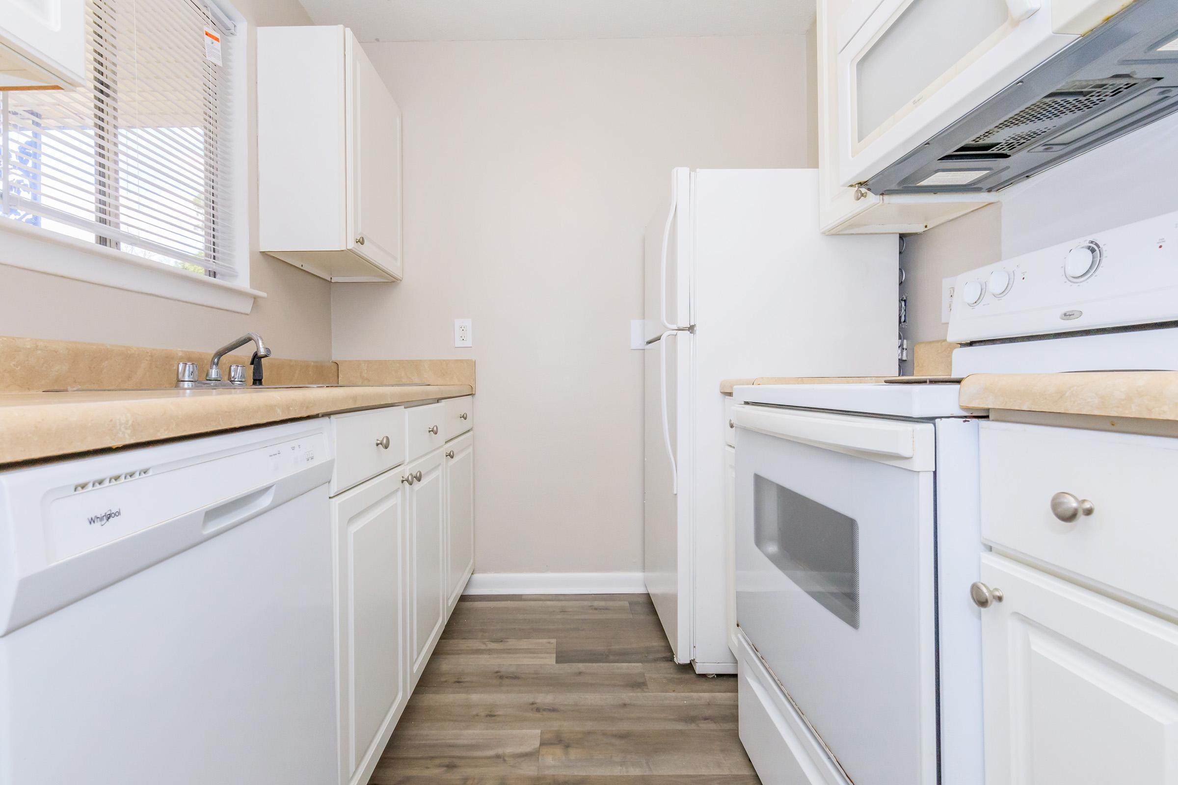 A modern kitchen featuring light-colored cabinets, a white refrigerator, a dishwasher, and an electric oven. The countertops are beige, and the flooring is a medium wood tone. Natural light is coming in through a window, creating a bright and inviting atmosphere.