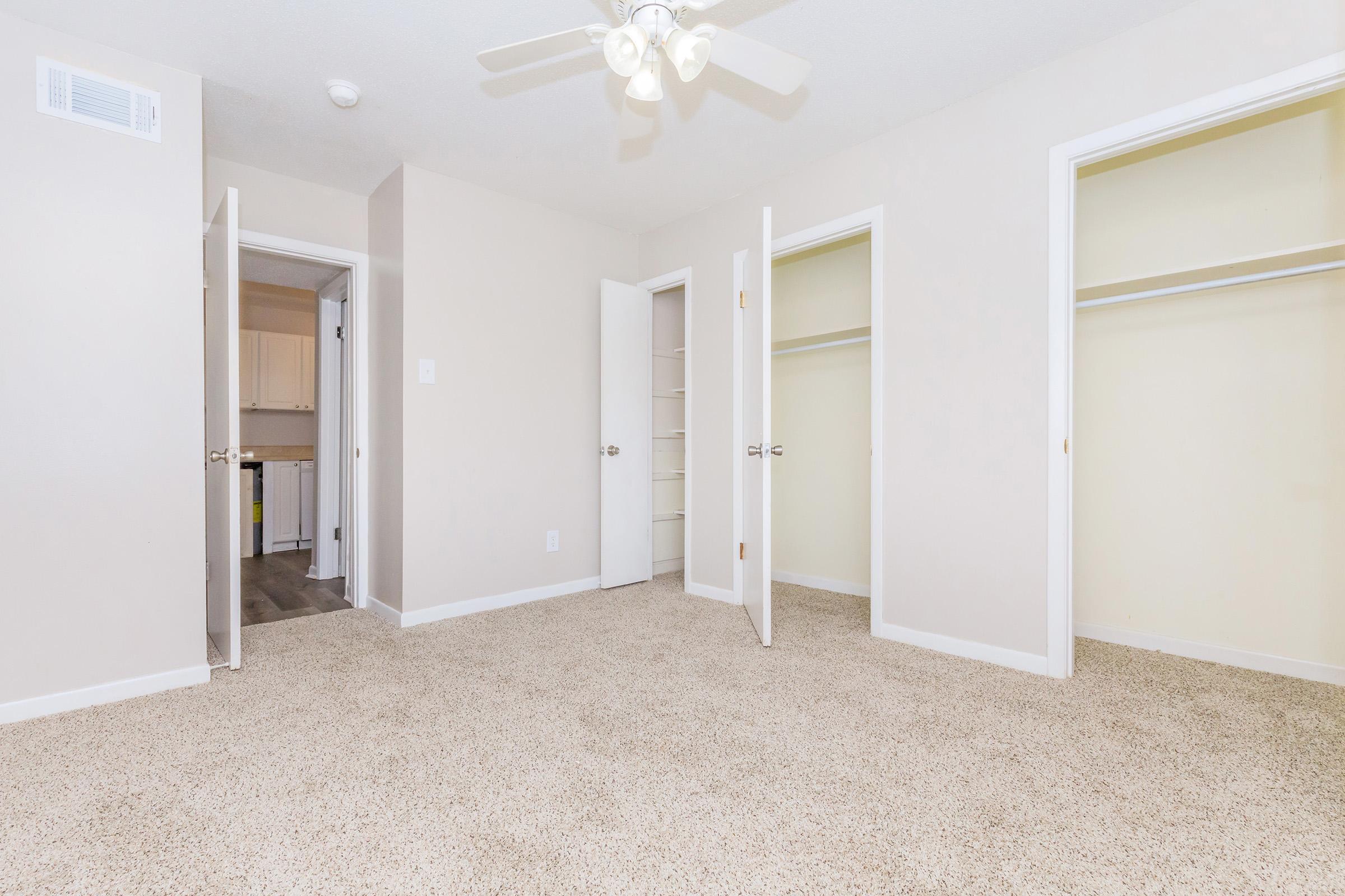 A spacious, empty room with light beige walls and carpet. There are two open doors leading to closets, and a ceiling fan above. Additional doors lead to different areas, providing a bright and airy feel to the space.