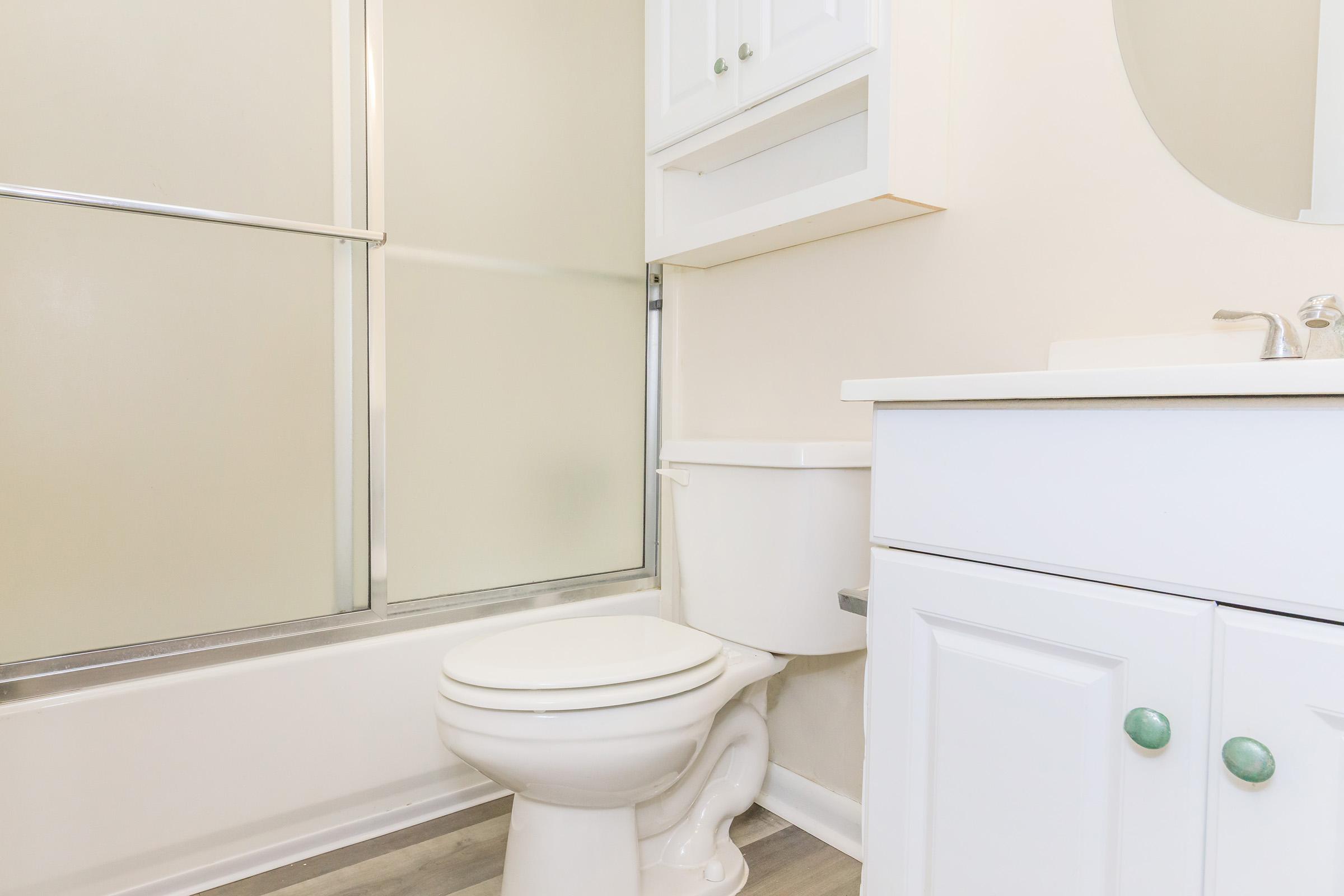 A clean and modern bathroom featuring a white toilet, a sink with a cabinet below, and a shower area with a glass door. The walls are painted light beige, and there is a round mirror above the sink. The floor has a light-colored wood finish, creating a bright and spacious appearance.