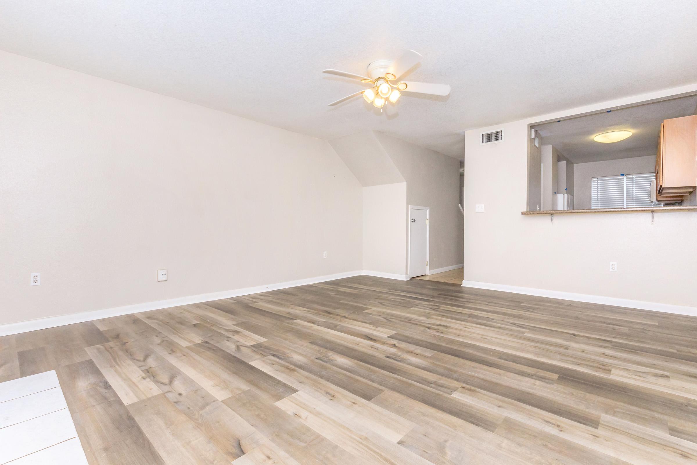 A spacious, well-lit living room featuring light wood flooring, a ceiling fan, and neutral-colored walls. There is an open view towards a kitchenette with wooden cabinets in the background, and a doorway leading to another room. The space appears empty, ready for furniture placement.