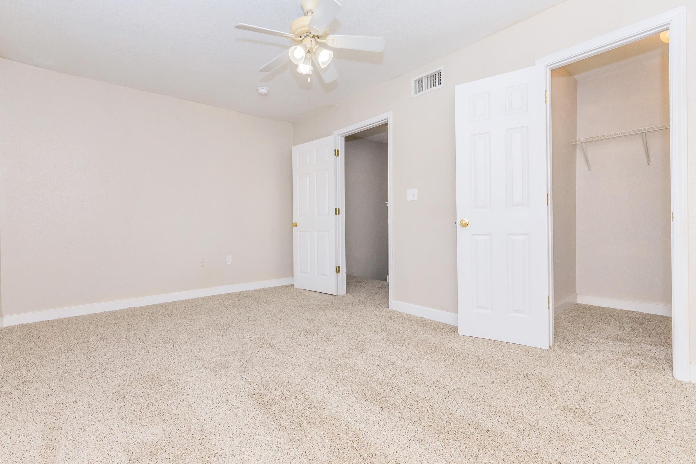 A spacious, empty room featuring beige walls, a ceiling fan, and light-colored carpeting. Two white doors are visible: one leading to a closet and the other to an adjacent room. The room has a bright and airy feel, with ample natural light.