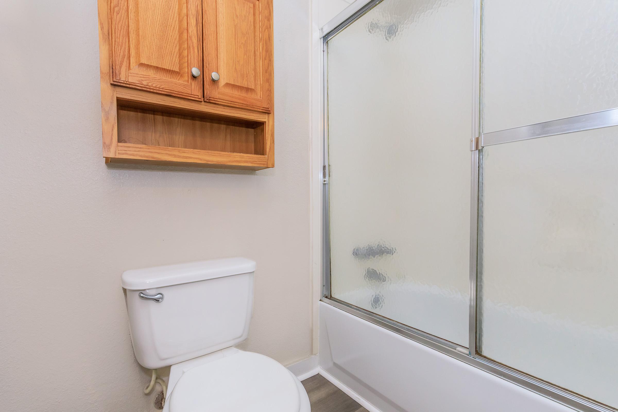 A small bathroom featuring a white toilet and a glass shower enclosure. There is a wooden cabinet above the toilet, and the walls are painted a light color. The bathroom floor has a neutral tile design.