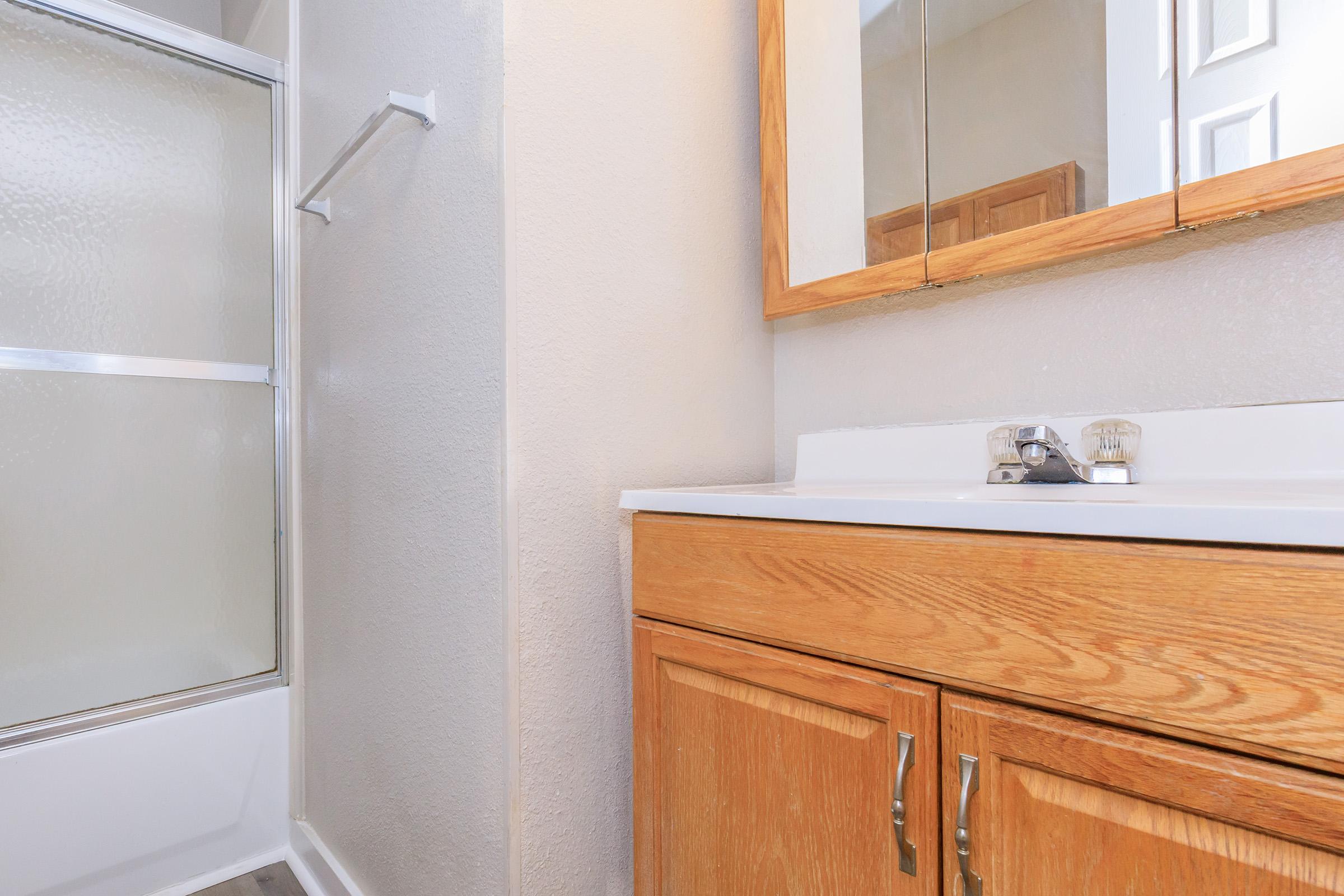 A bathroom scene featuring a shower stall with a glass door and a wooden vanity with a white countertop. The vanity has two wooden cabinets underneath. A mirror is mounted above the sink, reflecting part of the wall and the bathroom fixtures. The setting is well-lit and clean.