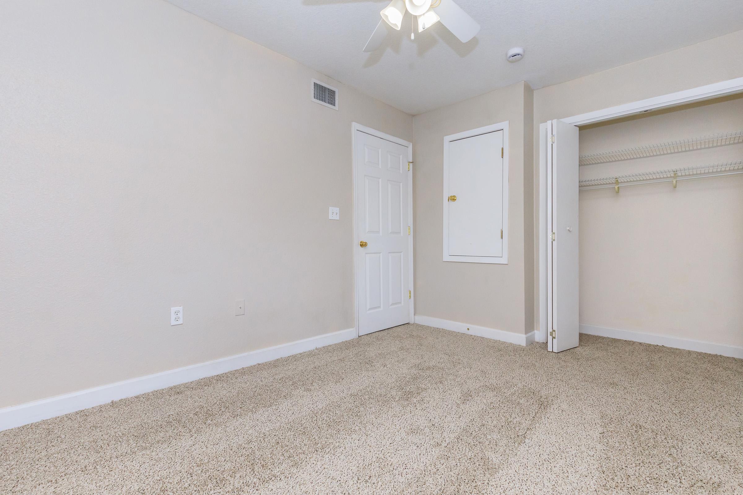 A clean, empty bedroom with light beige walls and carpet. It features a ceiling fan, white doors, and a small closet with a shelf. Natural light can be seen coming from the door and window, creating a bright and spacious atmosphere.