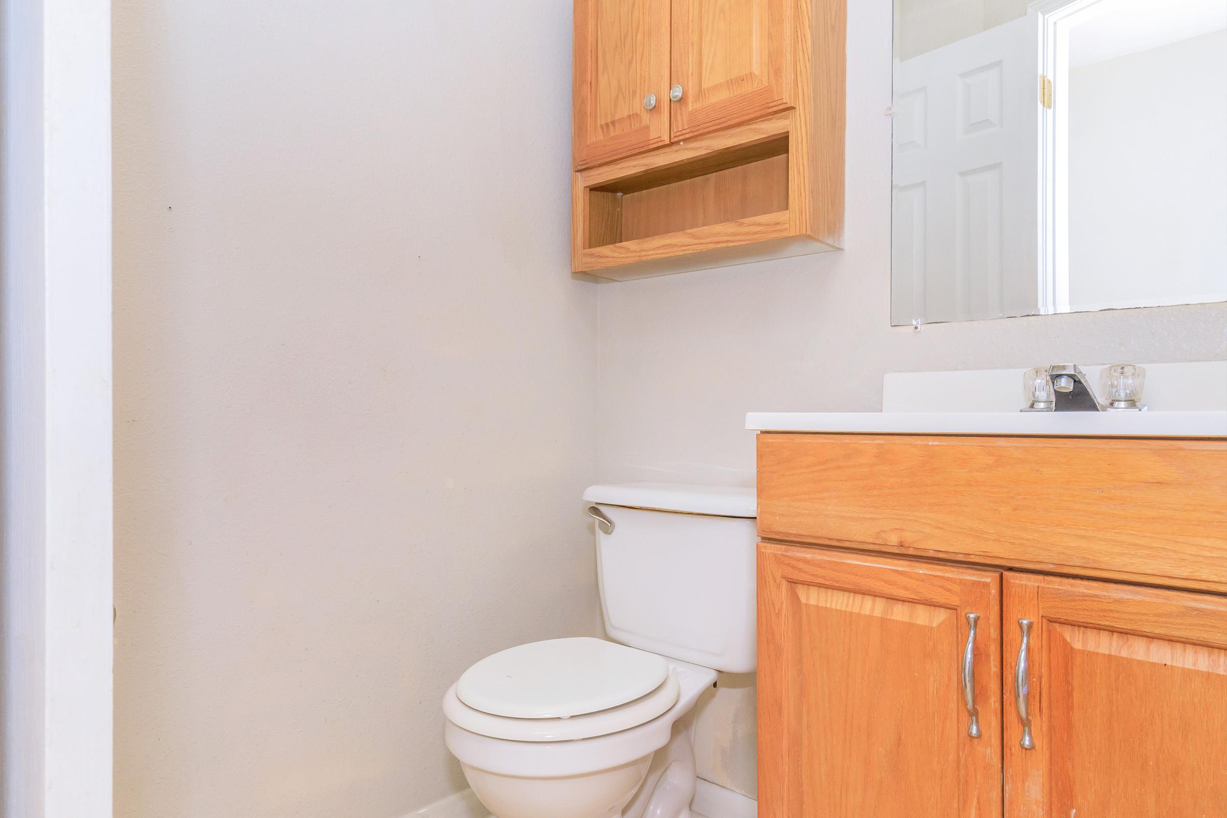 A small bathroom featuring a toilet, a wooden sink cabinet with drawers, and a mirror. The walls are painted a neutral color, and there is a shelf above the toilet. The floor is tiled, adding to the simple, functional design of the space.