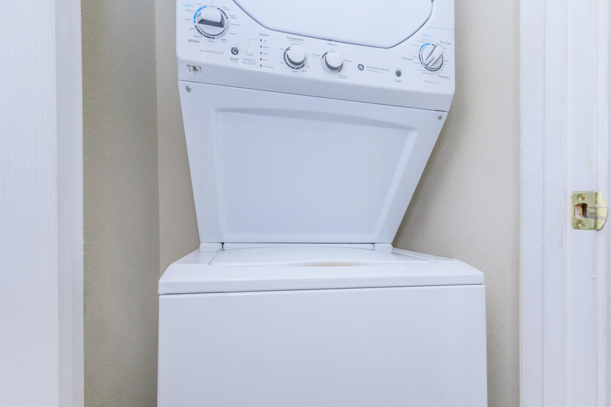 Stacked white washing machine and dryer in a narrow space, with control dials and buttons clearly visible on the front of the dryer. The wall behind is a neutral color, and the doors leading into the room are partially visible on either side.