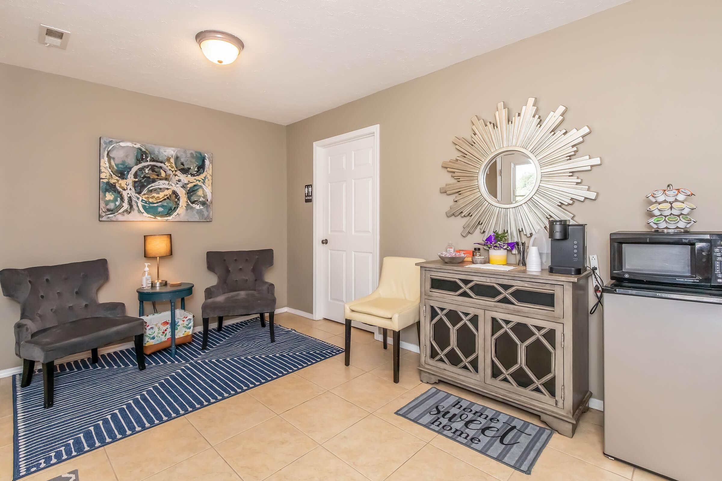 A cozy waiting area featuring two gray armchairs and a yellow chair, a small table with a lamp, a decorative wall piece, a mirrored sunburst, and a light-colored console table with coffee-making supplies and a microwave. The floor is tiled, and a striped rug adds to the decor.