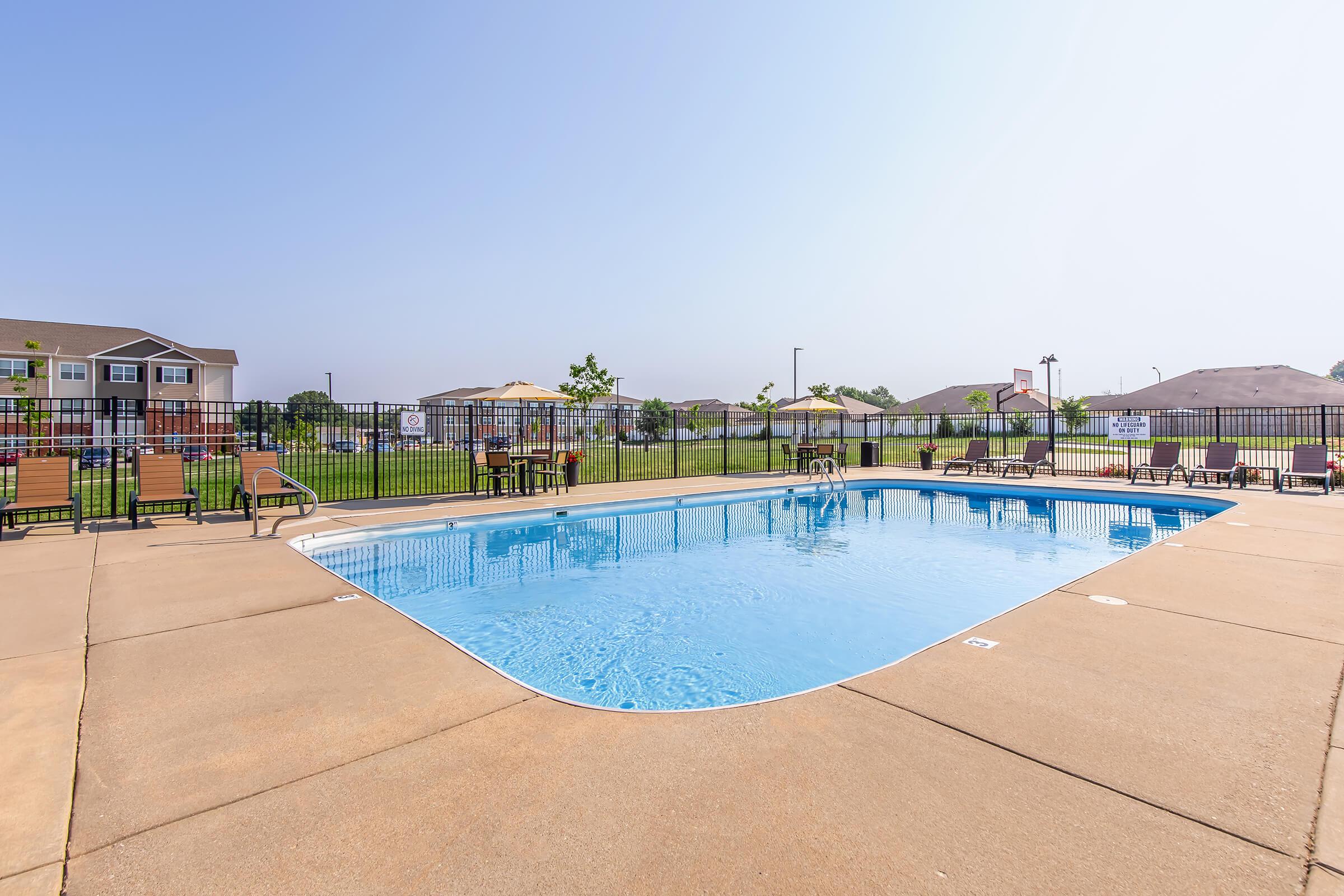 An outdoor swimming pool surrounded by a concrete deck, with lounge chairs and umbrellas. The pool is rectangular and has clear blue water. In the background, there are grassy areas and residential buildings. A fence encloses the pool area, providing privacy and safety.