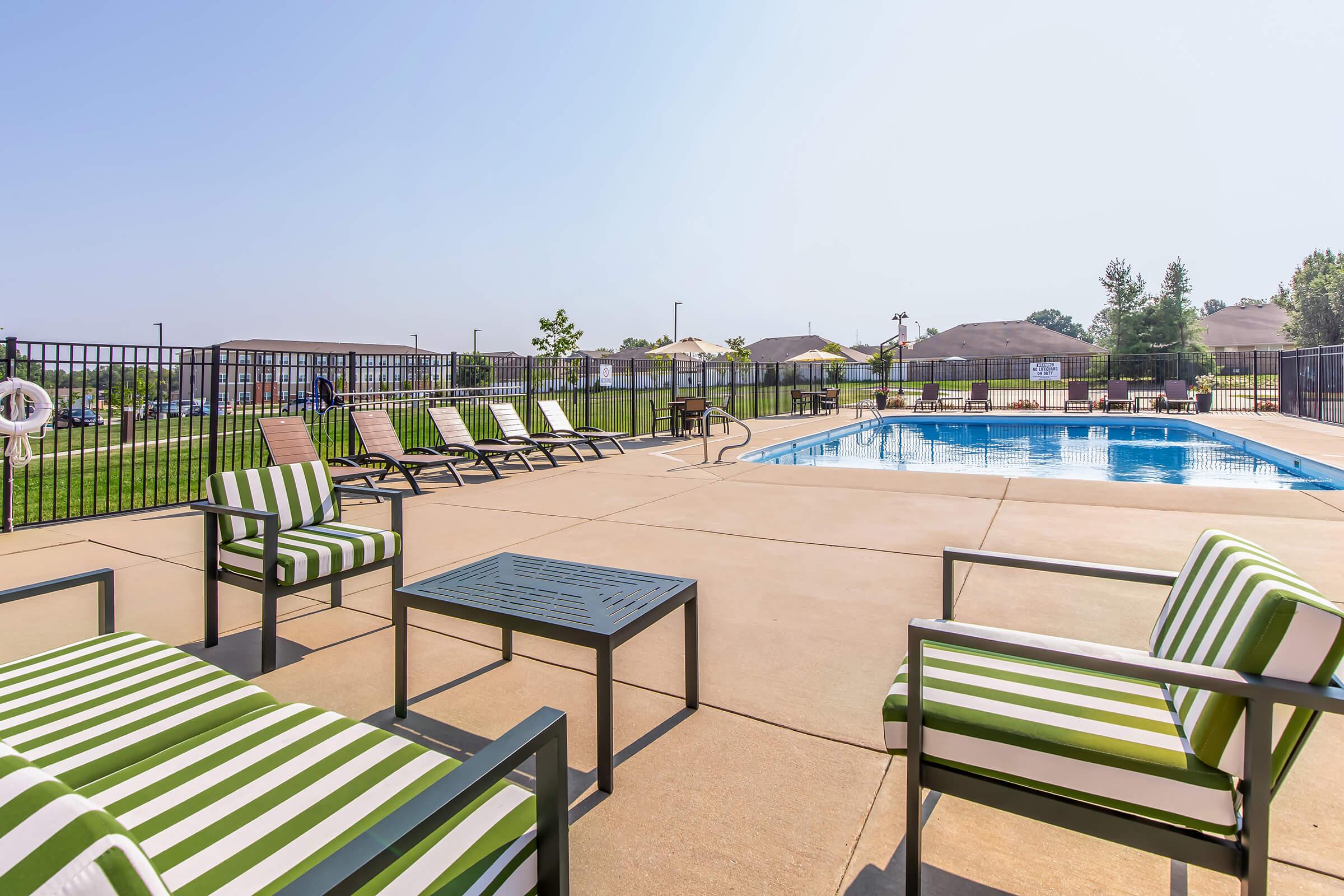 A spacious outdoor area featuring a swimming pool surrounded by lounge chairs with green and white stripes. There are several tables and chairs nearby, and the setting includes grassy areas and light residential buildings in the background under a clear blue sky.