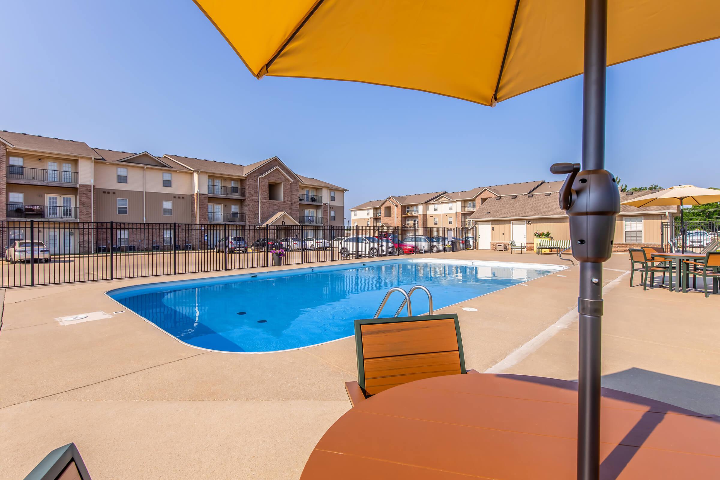 A sunny view of an outdoor swimming pool surrounded by lounge chairs and tables. An umbrella provides shade, and there are residential buildings in the background. The clear blue sky complements the inviting atmosphere of the pool area.