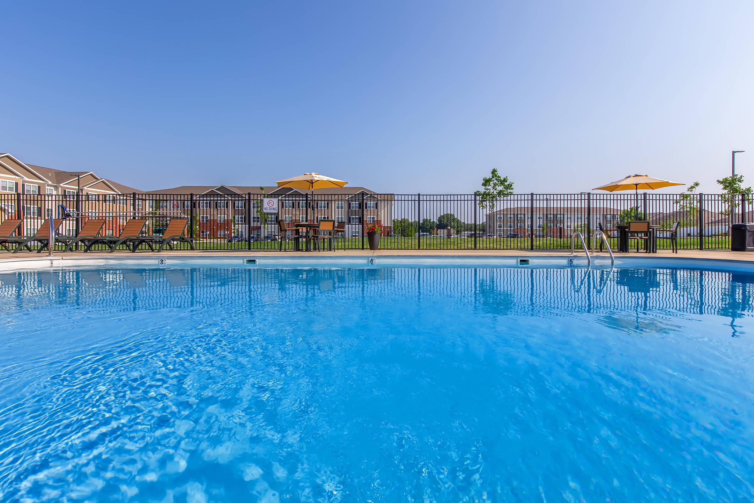 A clear blue swimming pool surrounded by lounge chairs and umbrellas, with a fence in the background. The setting features manicured grass and a few trees, under a bright blue sky. The atmosphere conveys a relaxed and inviting outdoor leisure area.