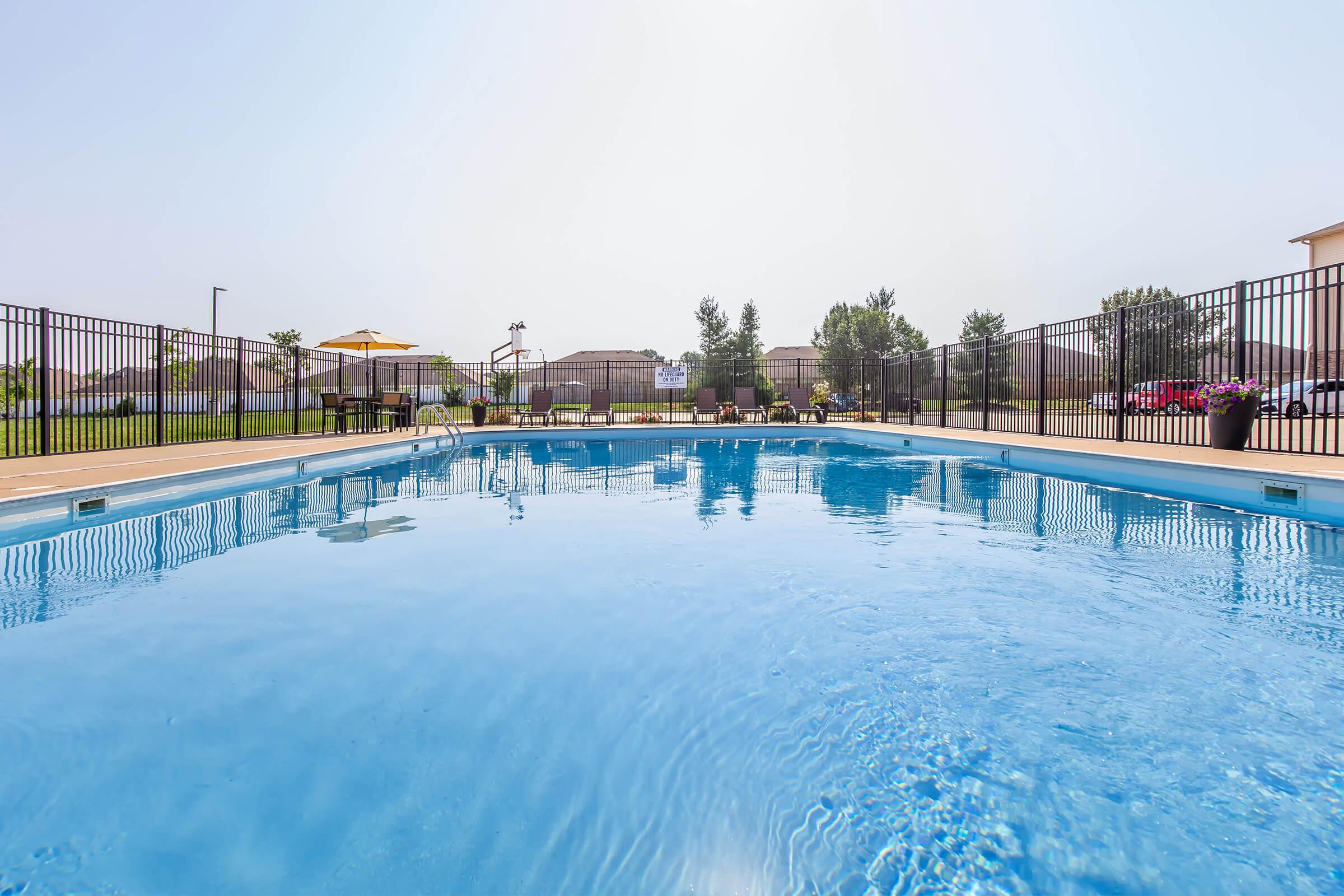 A clear, inviting swimming pool surrounded by a black metal fence. Sunlight reflects off the water's surface, and lounge chairs under umbrellas are visible in the background. Lush greenery and buildings are in the distance, indicating a well-maintained outdoor space.