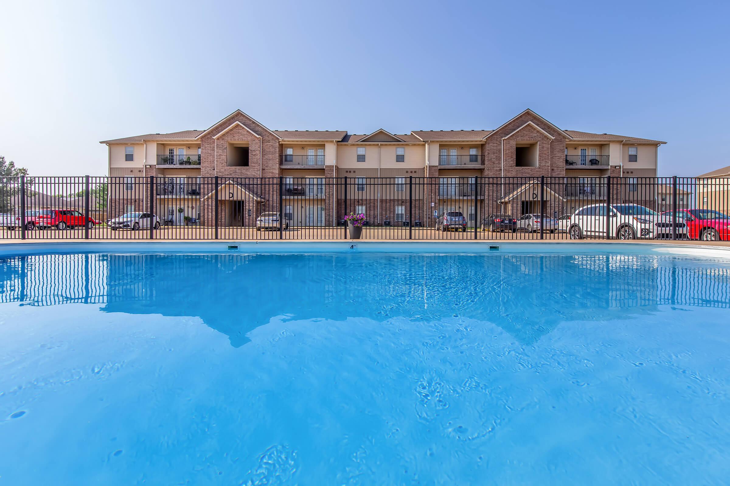 View of a residential area featuring a swimming pool in the foreground. The pool is clear and inviting, with a fence surrounding it. In the background, there are two three-story apartment buildings with balconies. Several cars are parked nearby, and the sky is clear and bright.