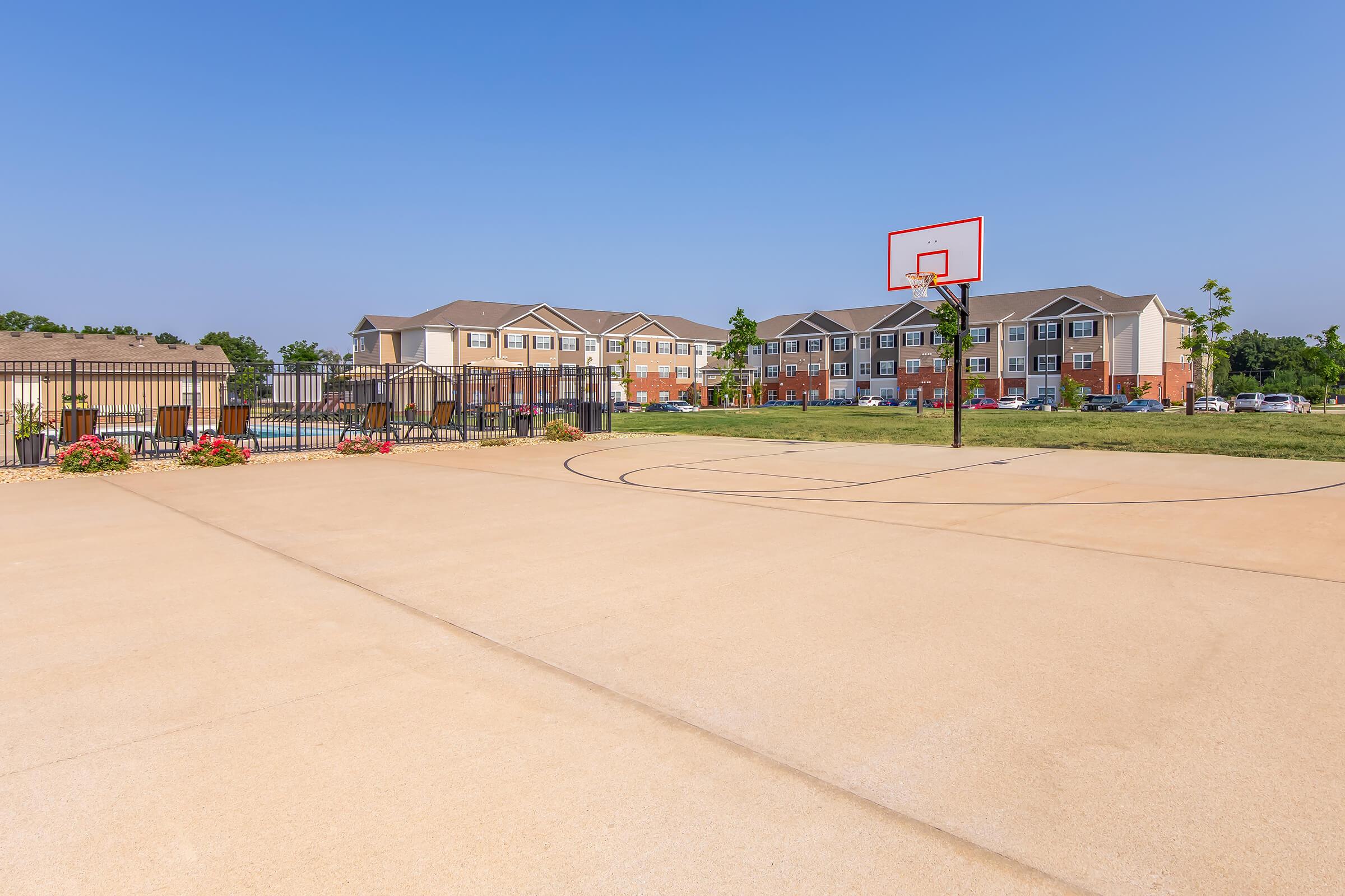 A basketball court with a hoop on the left side, surrounded by a fence and landscaped with flowers. In the background, there are residential buildings with multiple units, set against a clear blue sky. The court has a smooth concrete surface and outlines for the game.