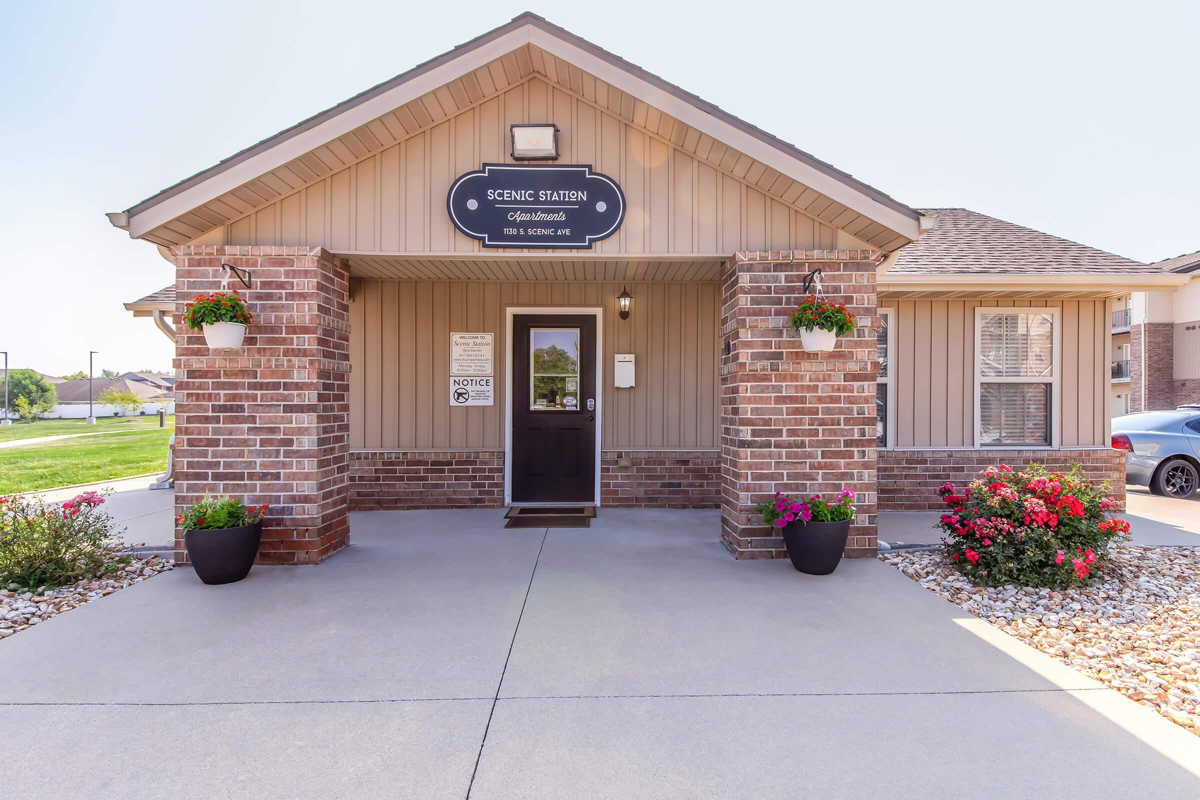 Exterior view of the Scenic Station Apartments entrance. The building features beige siding and a brick facade, with a covered porch, planters filled with colorful flowers on either side, and a sign above the door. A notice is visible near the entrance, and a car can be seen parked nearby.