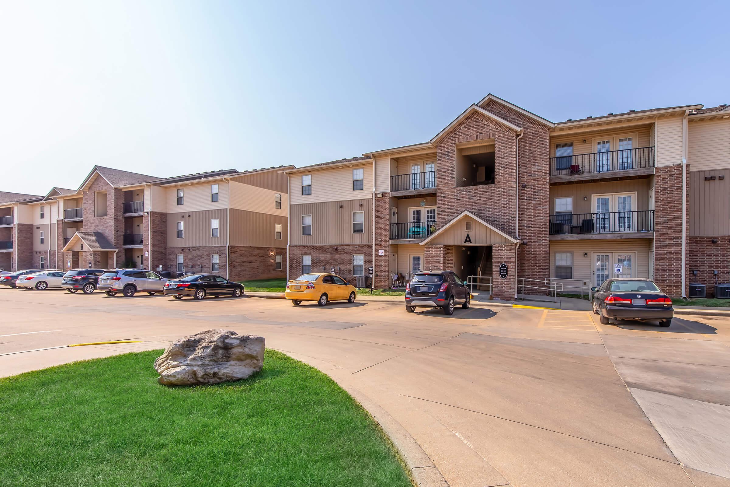 A view of a residential apartment complex featuring multiple buildings with brick and siding exteriors. Cars are parked in front of the buildings, and there is a landscaped area with a large rock and green grass in the foreground. The sky is clear and blue, indicating a sunny day.