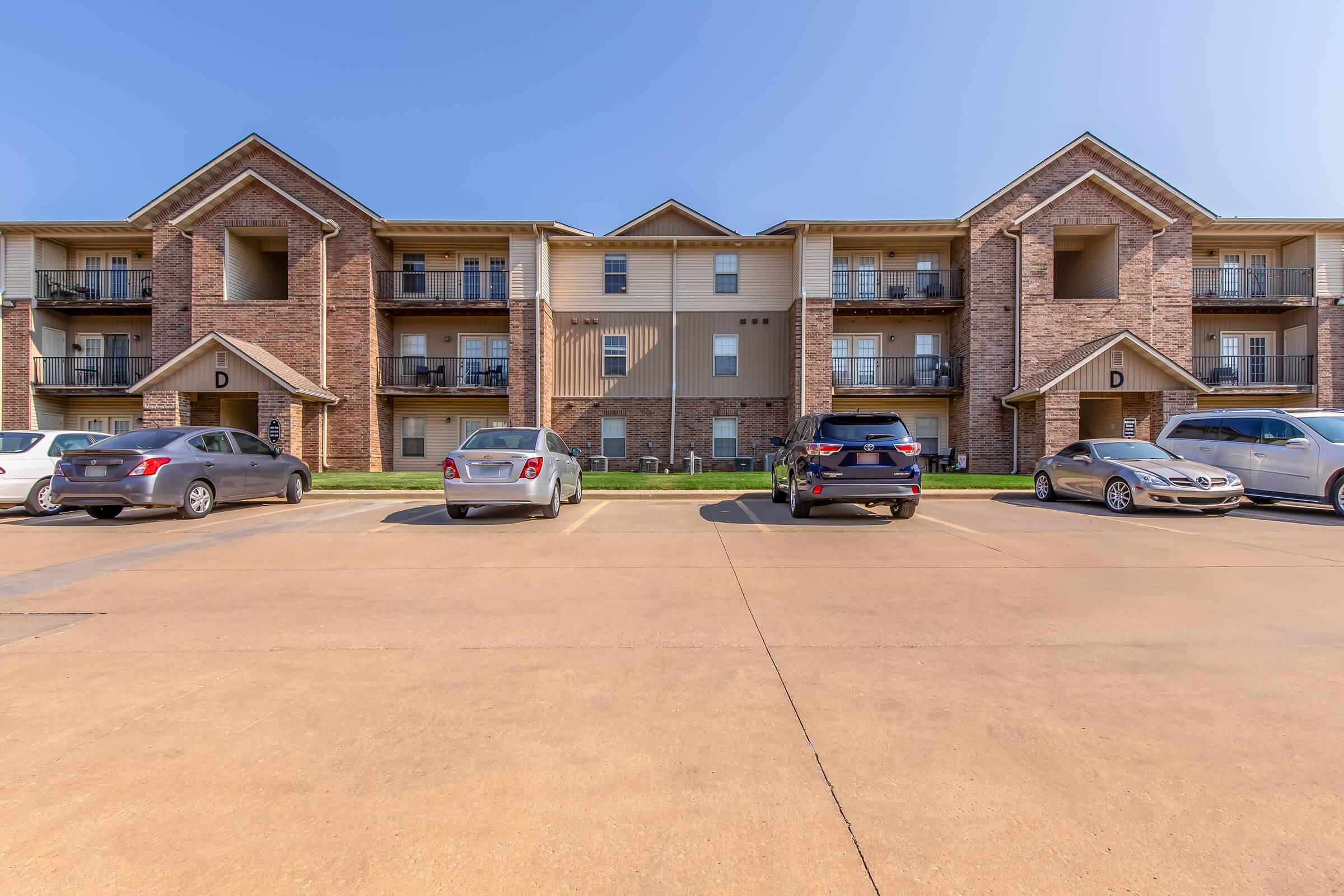 A row of multi-story apartment buildings with brick exteriors, featuring balconies on the upper floors. A paved parking area in front has several cars parked, including a black SUV, a silver sedan, and a gray vehicle. The sky is clear and blue, creating a bright, sunny atmosphere.