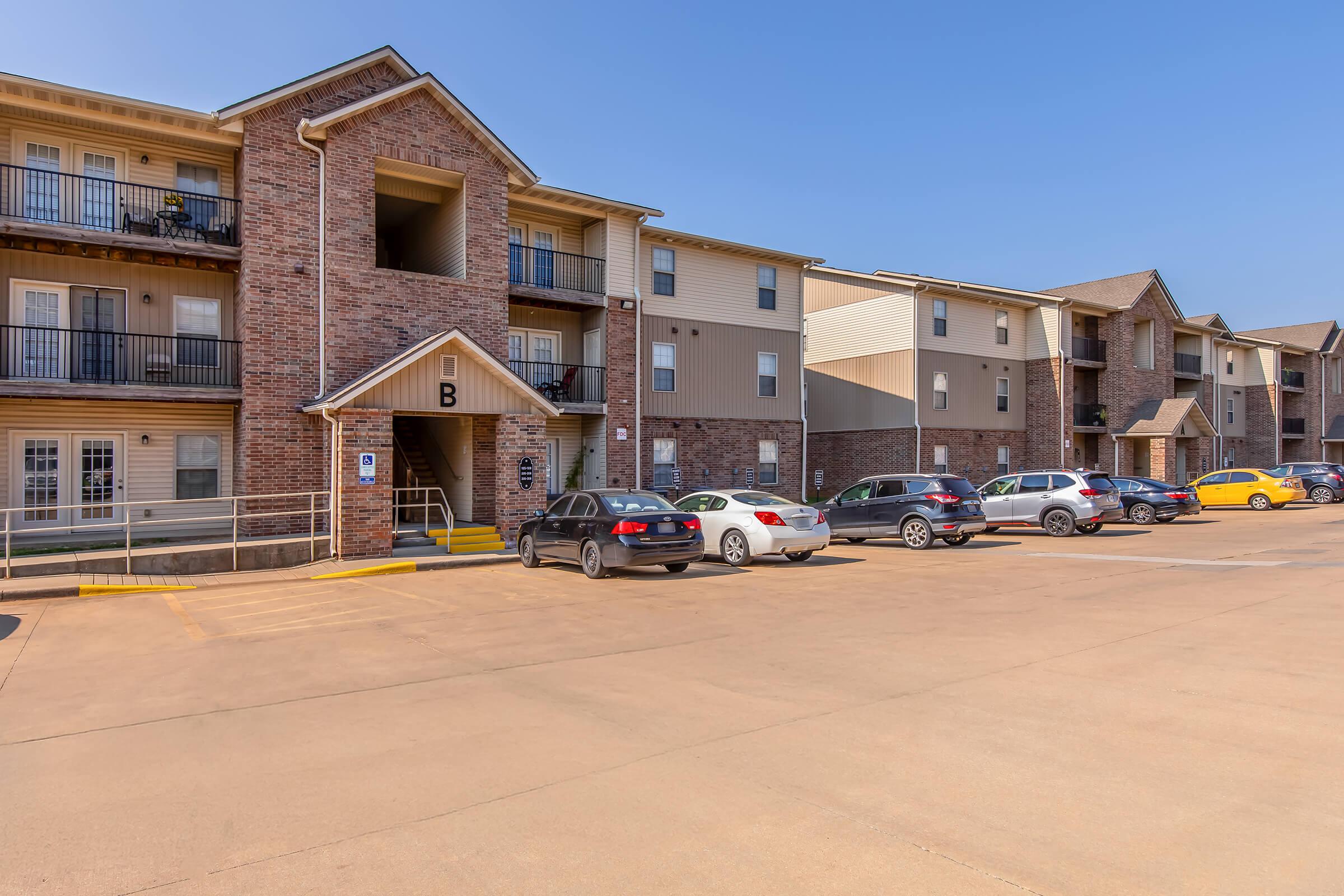 A three-story apartment building with brick and siding facade. The building features multiple balconies and a central entryway labeled "B." A parking lot is visible in front, filled with several parked cars. Clear blue sky overhead.