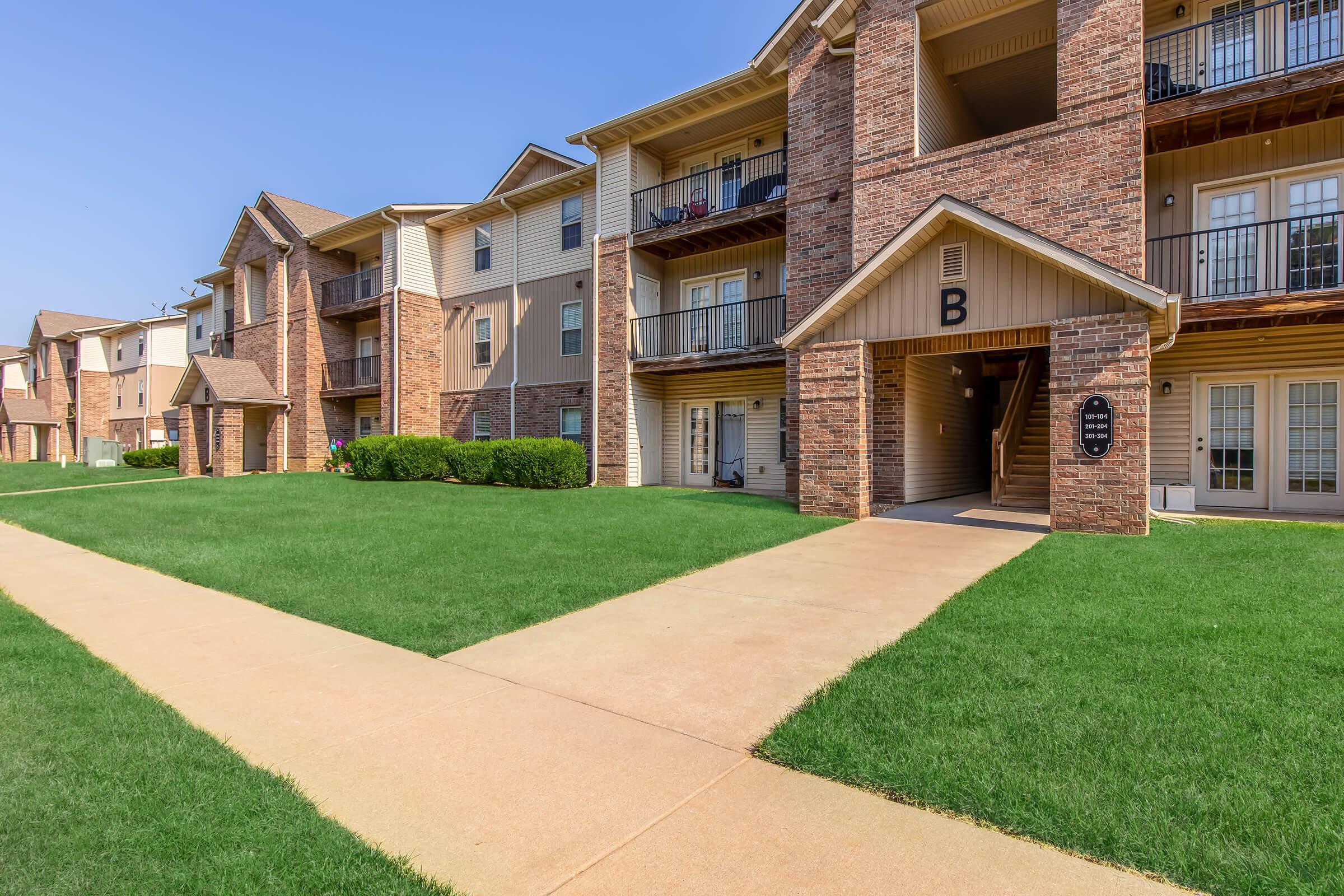 Exterior view of a multi-unit apartment building featuring brick and siding architecture. The image shows a well-maintained lawn and walkways leading to the entrance, with clear blue skies in the background. The building has multiple balconies and a distinct letter "B" marking one of the entrances.