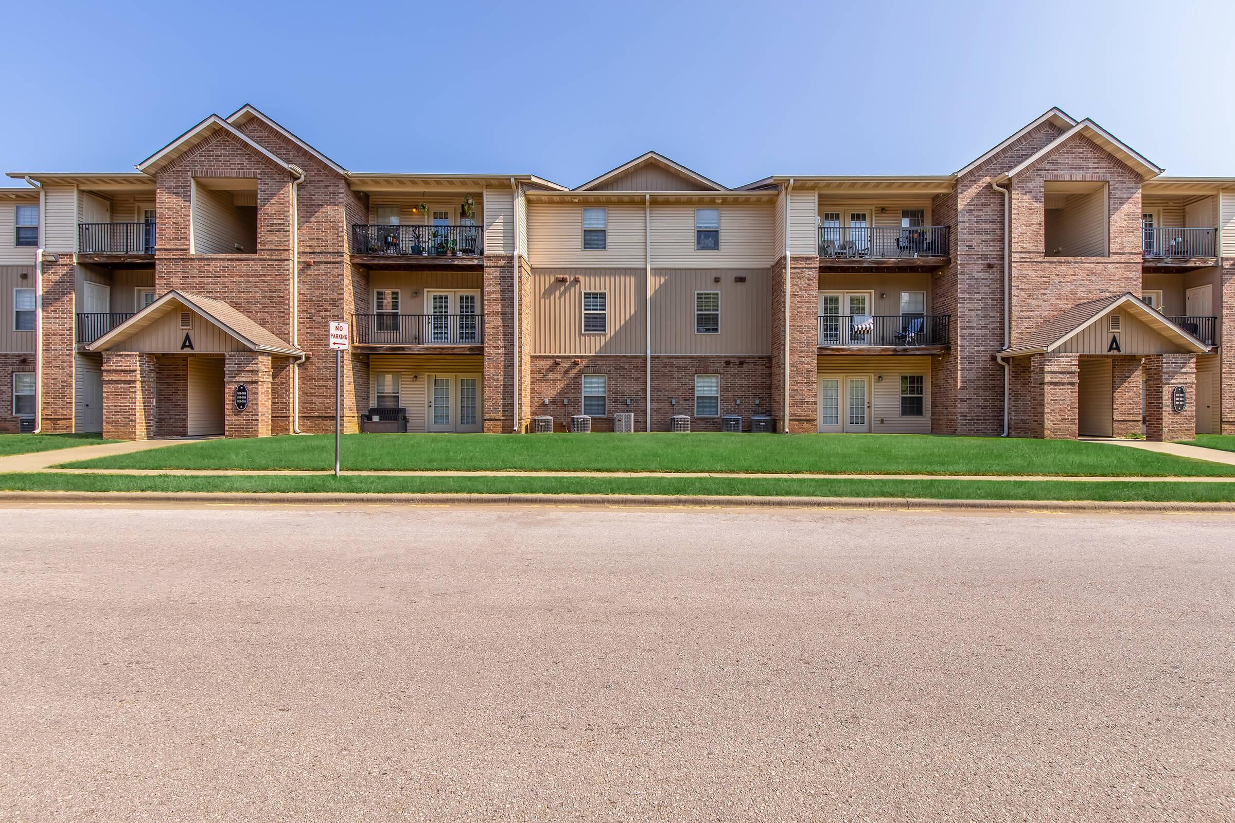 A multi-unit residential building featuring brick and beige siding. The two-story structure has balconies, with green grass in the foreground and a paved area leading to the entrance. The buildings are situated under a clear blue sky.