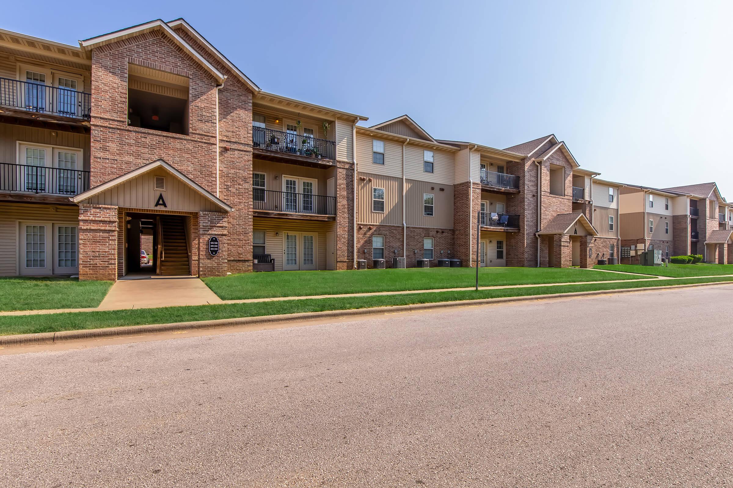 A row of residential apartments featuring brick and siding exteriors, balconies, and well-maintained lawns alongside a paved street under a clear sky. The buildings are two to three stories high, with some units displaying outdoor furniture on the balconies.