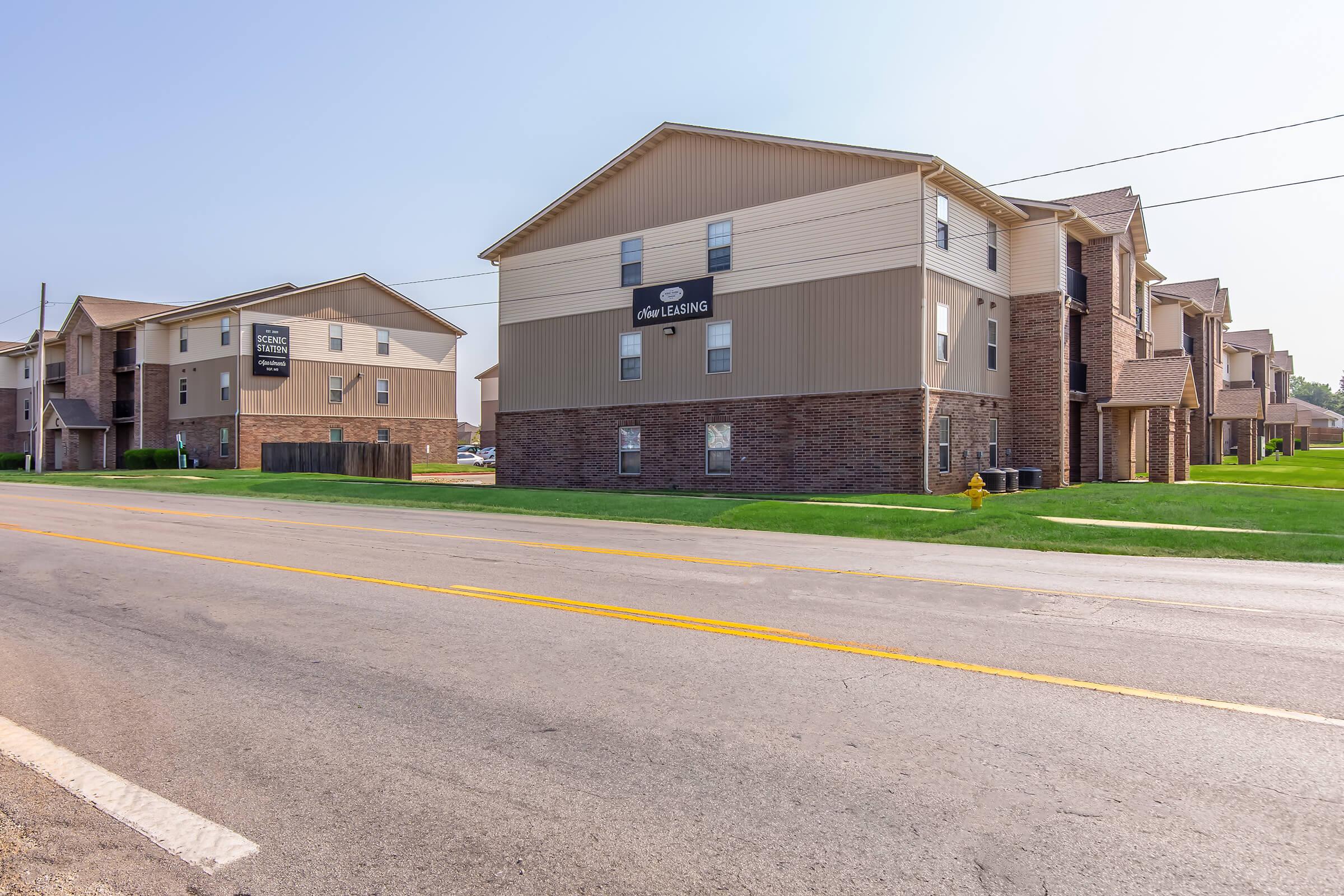 A view of two residential apartment buildings alongside a paved road, featuring grass lawns and clear blue skies. The buildings have a combination of brick and siding exteriors, with leasing signs on display, indicating availability. Street visibility shows a well-maintained area.
