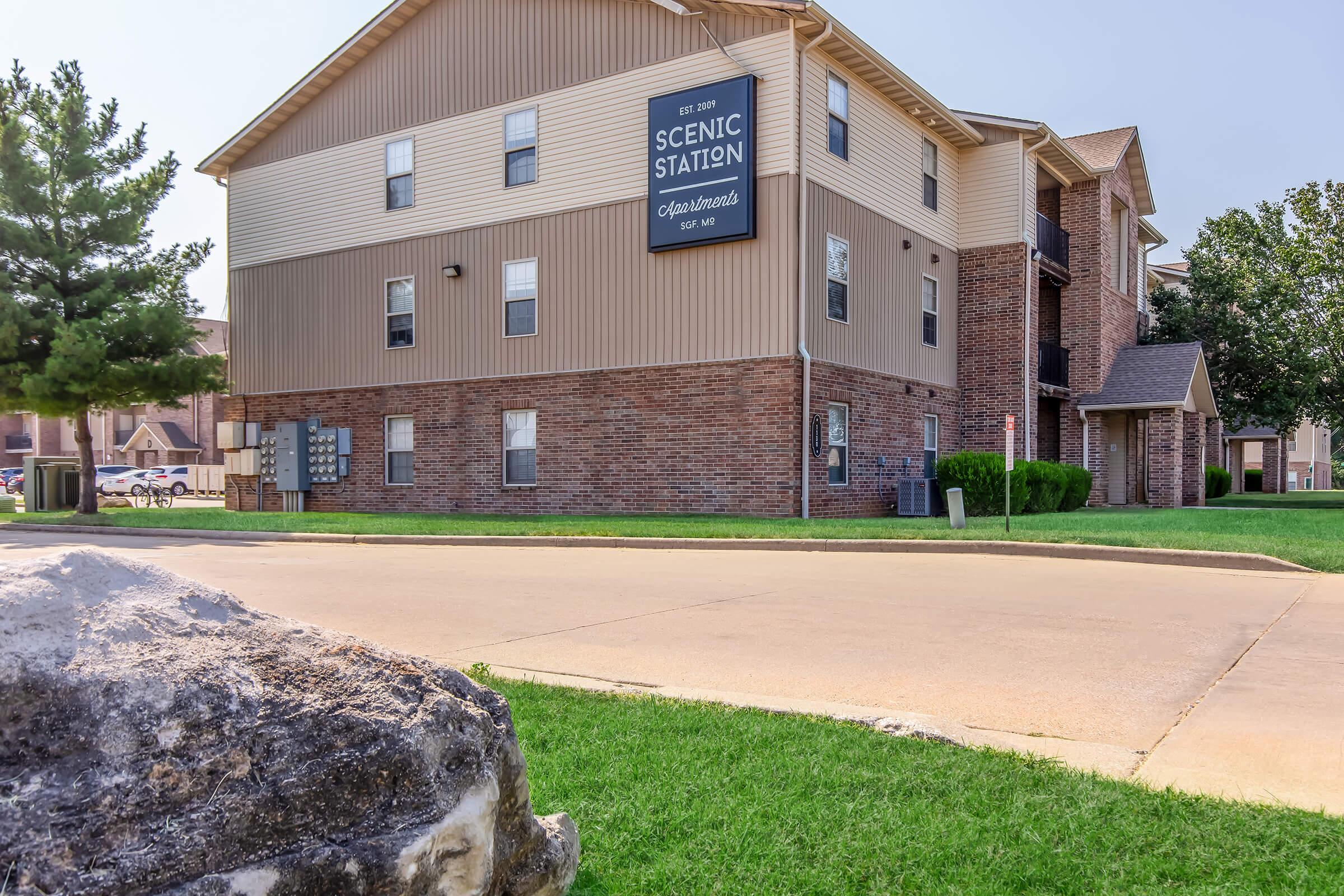 Exterior view of Scenic Station, a multi-story residential building featuring a mix of brick and siding architecture. The sign indicates the building's name. There is a grassy area in front, a large rock, and several utility boxes on the side. The sky is clear and blue.