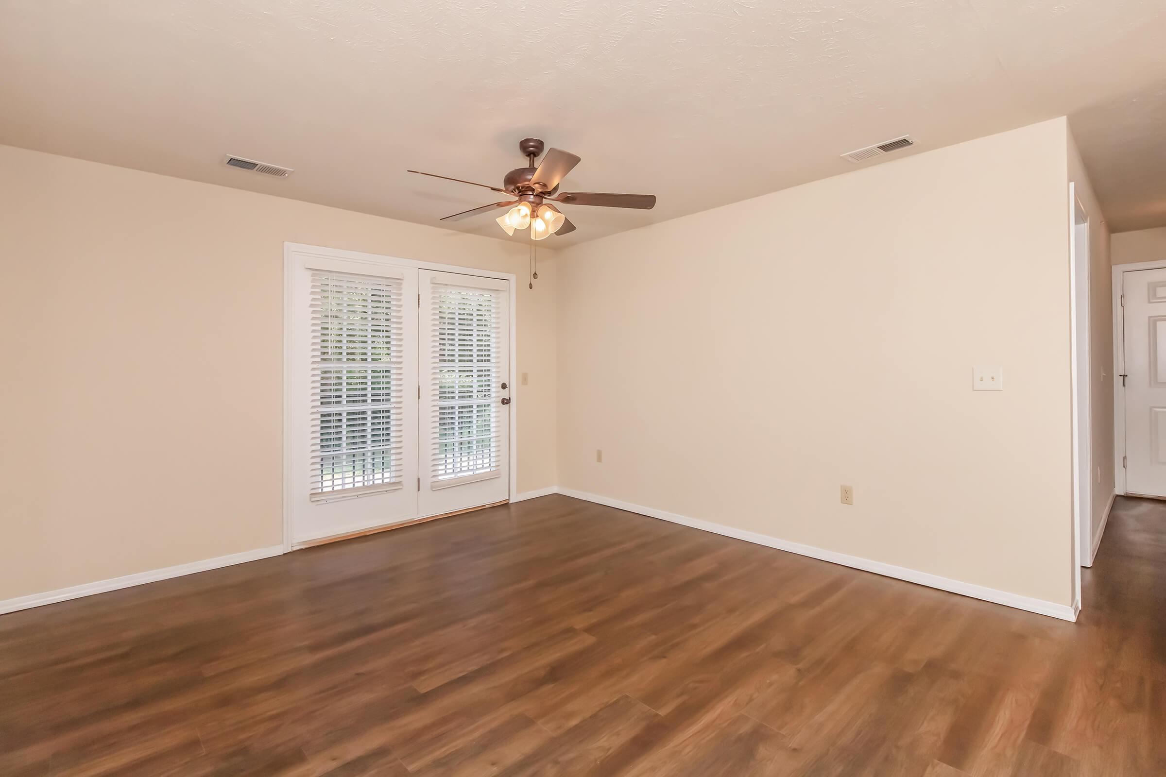 A spacious living room with light-colored walls and wooden flooring. There is a ceiling fan above and a set of sliding glass doors leading outside, partially covered by white window shutters. The room is empty, creating a bright and open atmosphere.