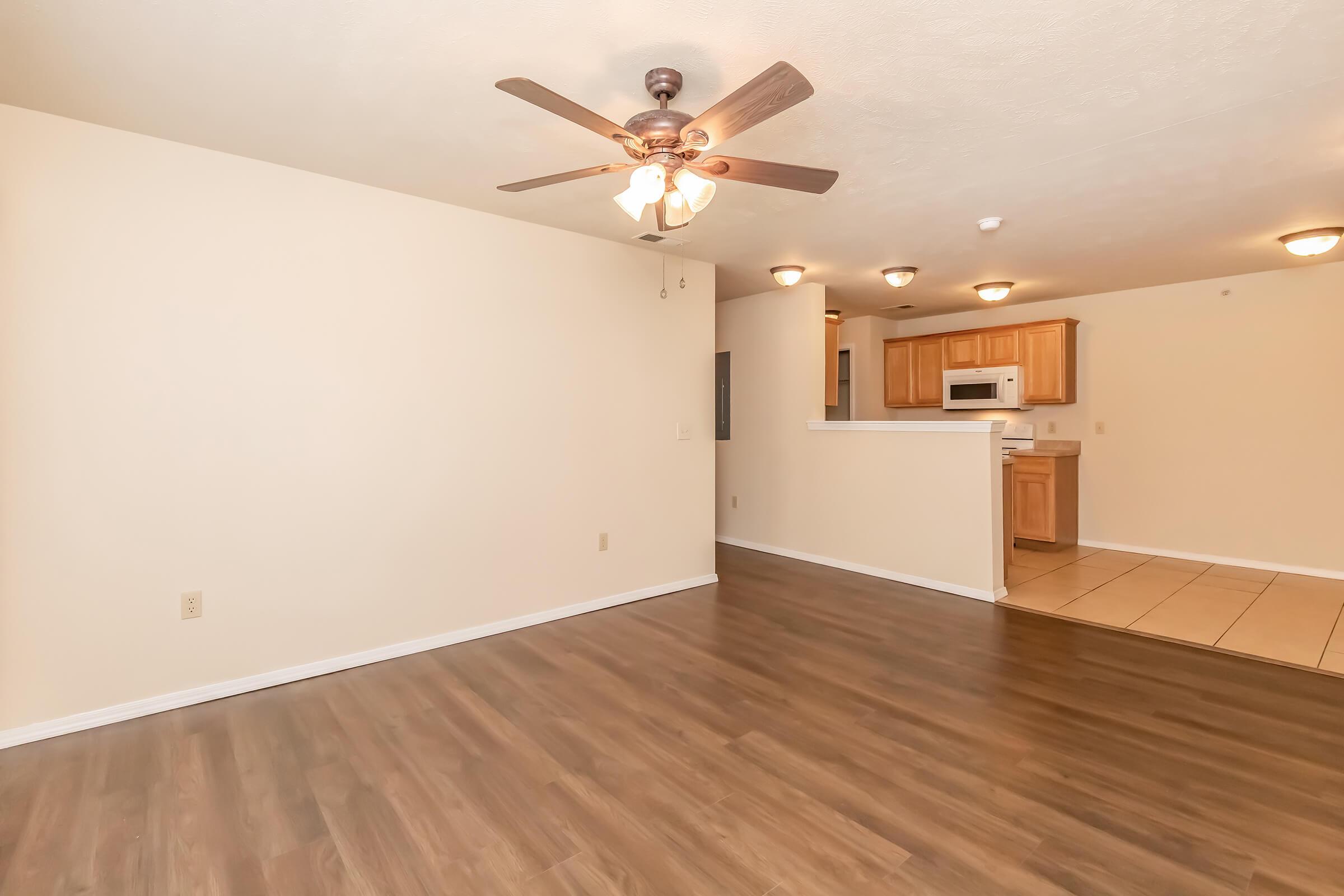A spacious, well-lit living area featuring hardwood flooring, a ceiling fan, and neutral-colored walls. In the background, there's an open kitchen with wooden cabinets, a microwave, and tile flooring. The layout suggests a modern and inviting home environment.
