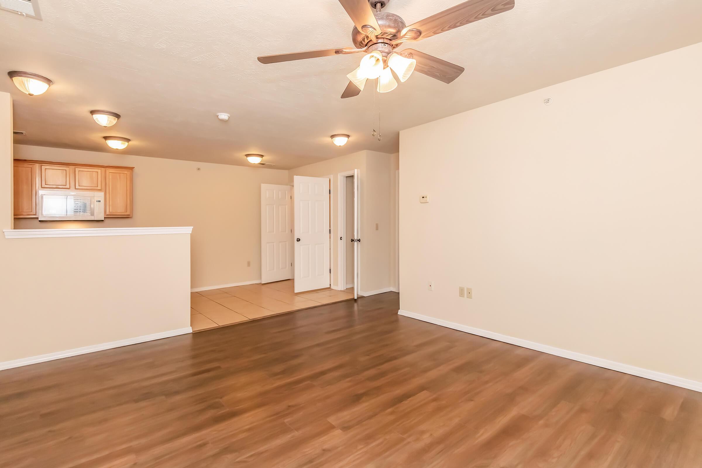 Interior view of an apartment featuring a wood laminate floor, ceiling fan, and recessed lighting. The space includes a kitchen area with light wood cabinets and a doorway leading to another room or hallway. The walls are painted in a neutral tone, creating a bright and open atmosphere.