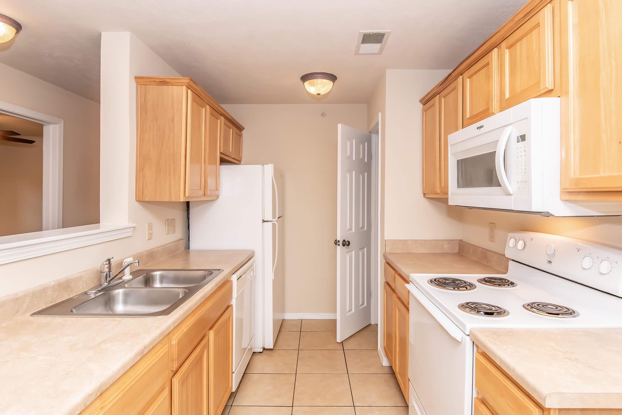 A modern kitchen featuring light wooden cabinetry, a double sink, white appliances including a refrigerator and microwave, and a stove. The walls are painted a light beige, and there are neutral-colored tiles on the floor. A door leads to an adjacent room, enhancing the open feel of the space.