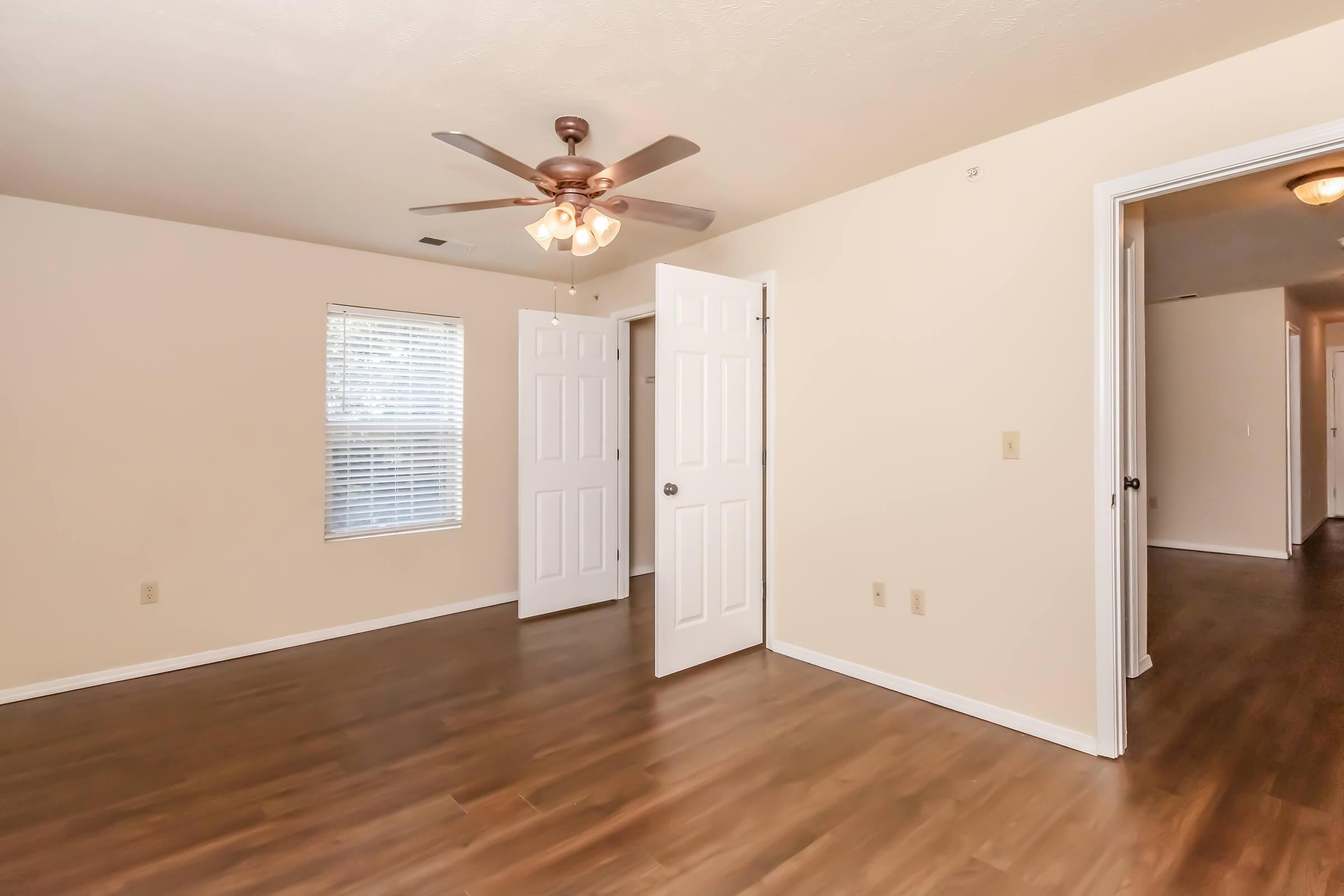 A well-lit room with light-colored walls and wooden flooring. It features a ceiling fan, two white doors, and a window with blinds. The room has a simple, empty layout, creating a spacious feel, with an additional door leading to another area.