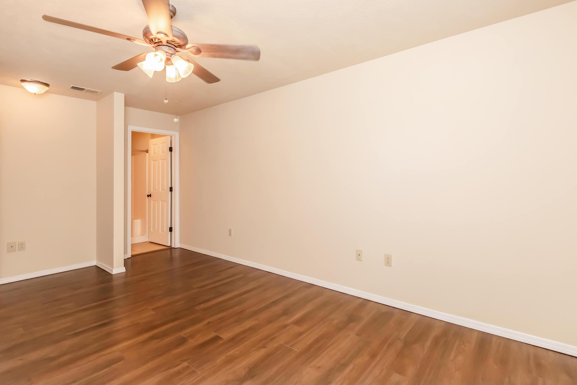 Empty room with light-colored walls and a ceiling fan. There is a doorway leading to another area, likely a bathroom, and the floor is made of wood laminate. The space is well-lit and spacious, providing a clean, minimalist look.