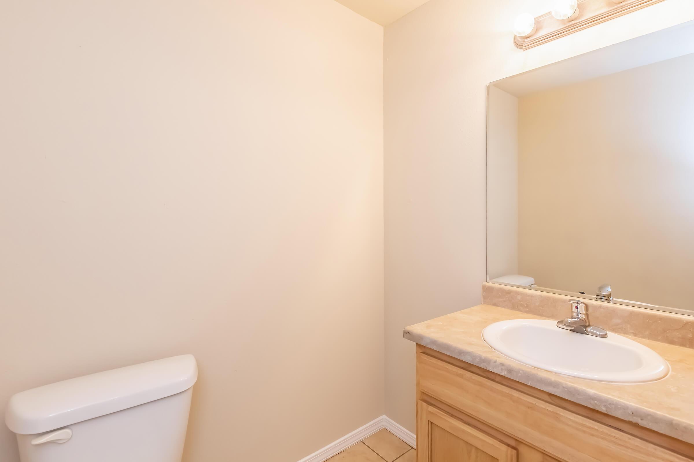 A simple bathroom featuring a light-colored wall, a countertop with a sink, a mirror above the sink, and a white toilet. The floor is tiled, and there is soft lighting from an overhead fixture. The space appears clean and well-maintained with minimal decor.