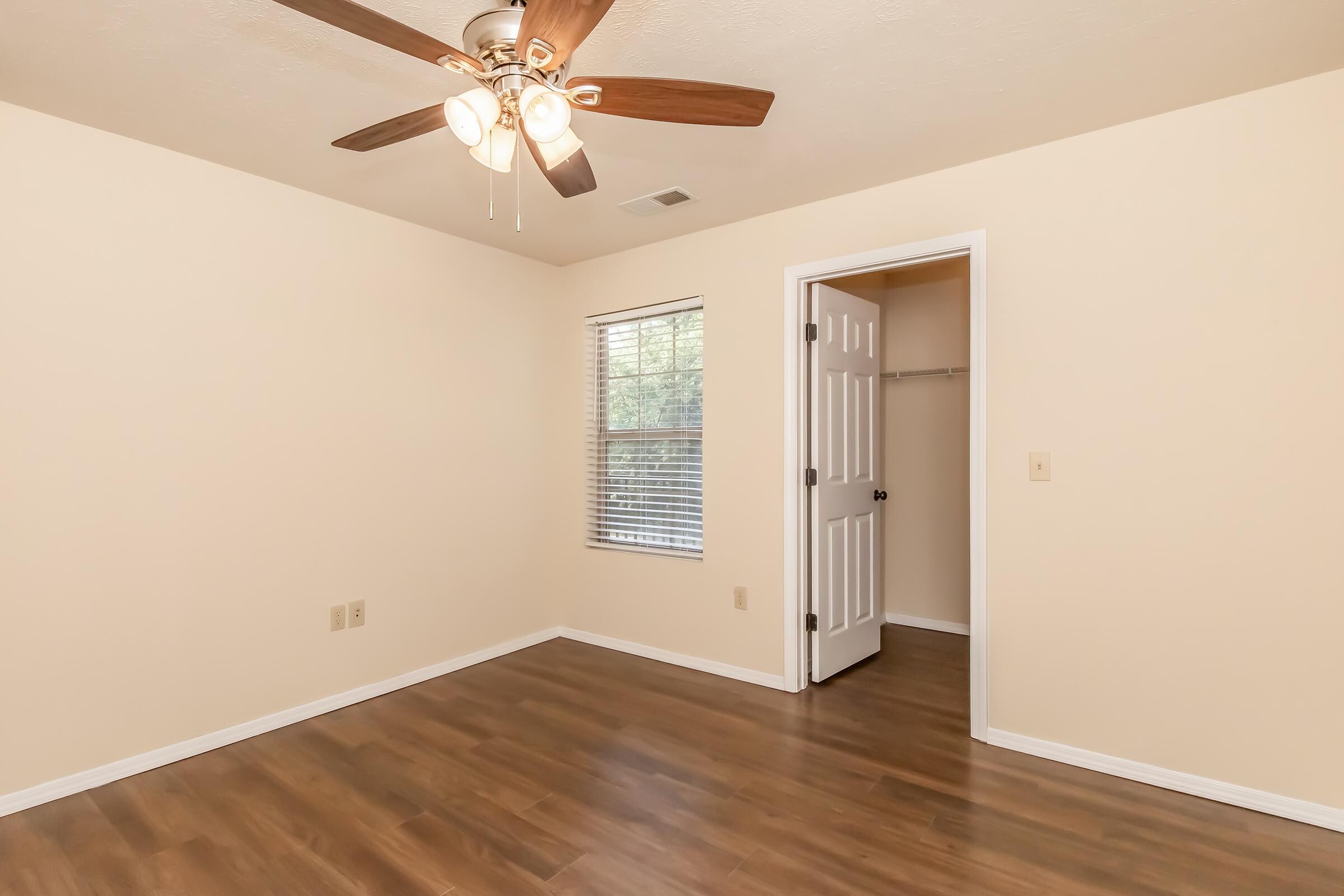 Bright and spacious bedroom featuring a ceiling fan, a window with blinds, and light-colored walls. A door leads to a closet area on the right, and the floor is finished with wood laminate, creating a warm and inviting atmosphere.