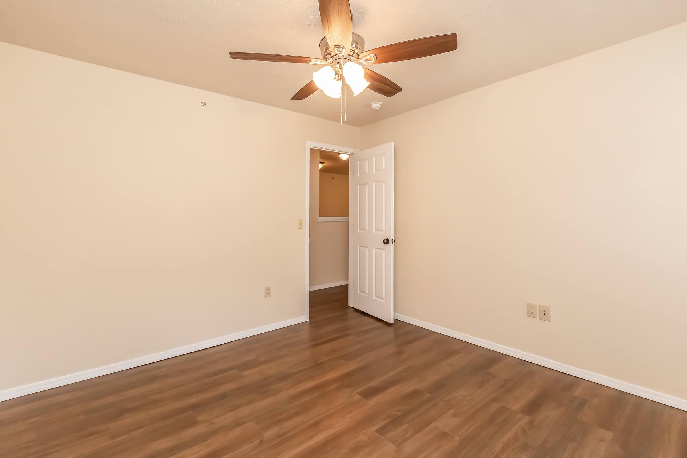 A bright, empty room featuring light-colored walls and a wood floor. A ceiling fan with wooden blades hangs from the ceiling. A white door is slightly open, revealing another room behind it. The space appears clean and well-lit, ideal for various uses.