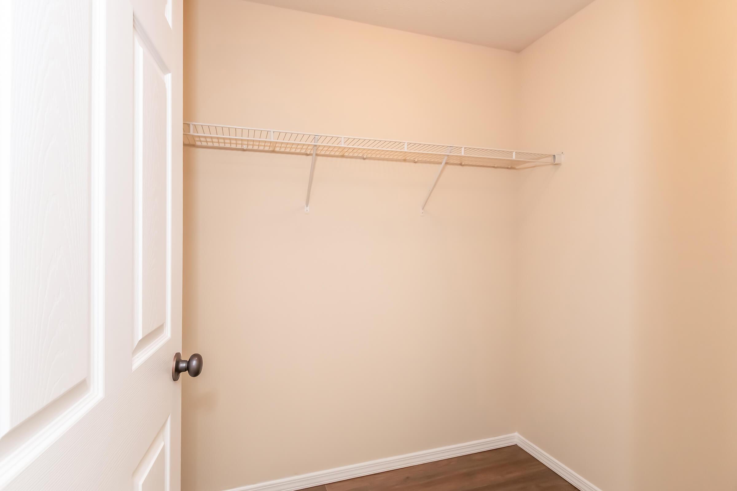 A clean, empty closet with beige walls and a ceiling-mounted wire shelf. The door is partially closed, revealing the spacious interior with no items stored inside. The floor is wood laminate, complementing the simple, minimalistic design of the space.