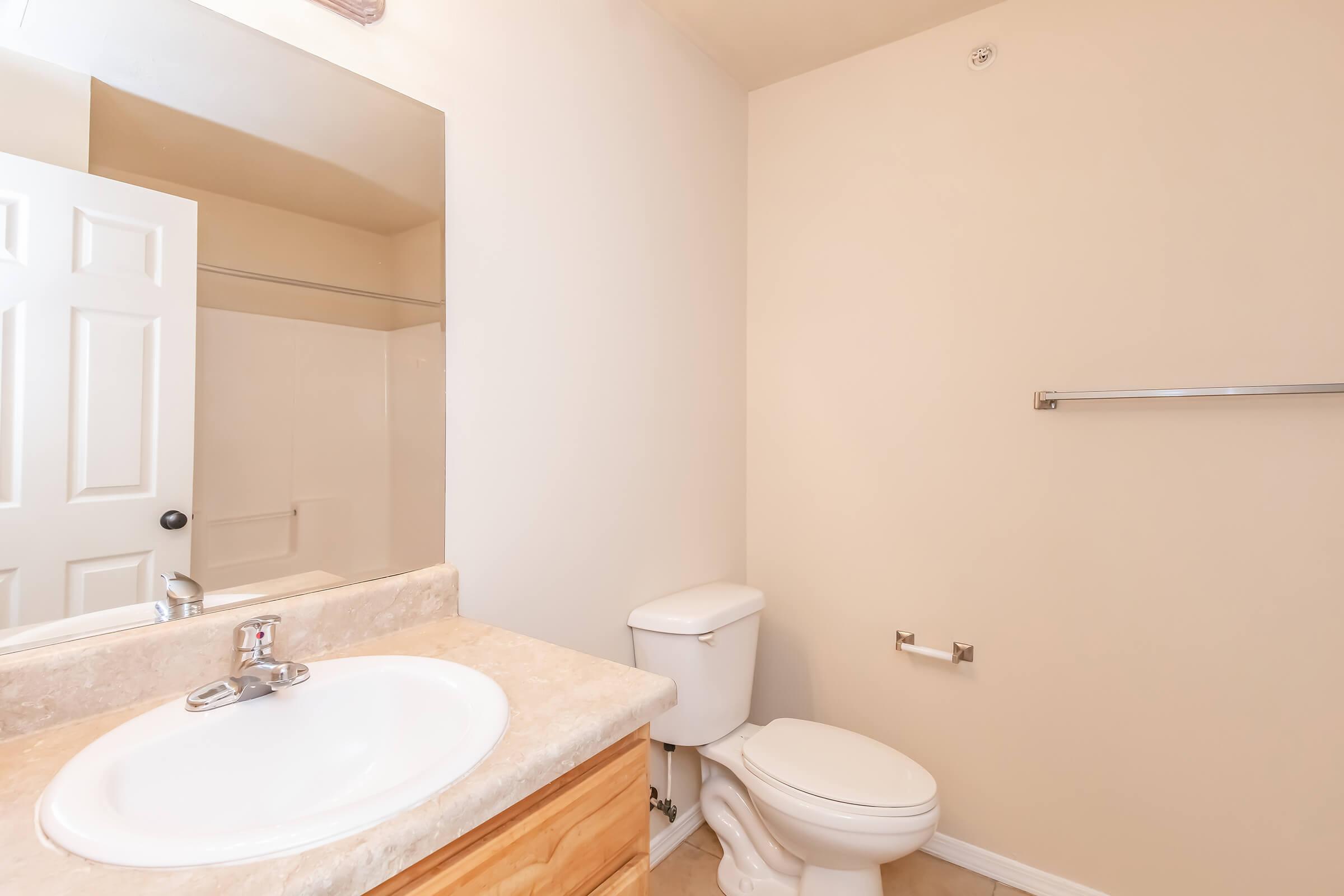 A simple bathroom featuring a beige wall, light-colored countertop with a sink, a mirror above the sink, a toilet, and a towel rack. A shower area is visible, and the door is partially open, revealing a closet space. The overall decor is minimal and clean.