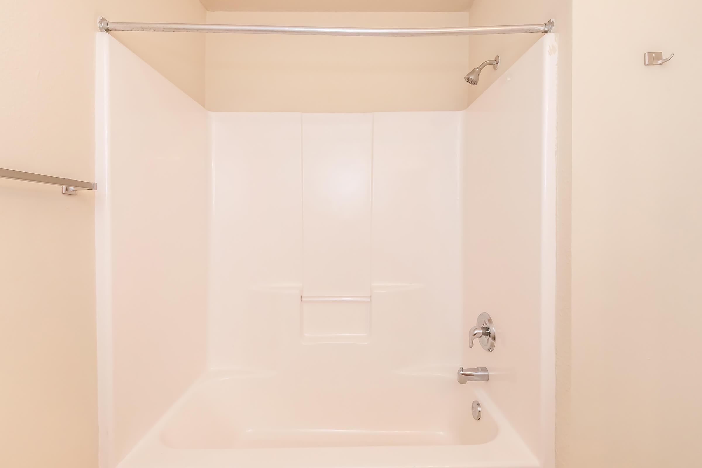 A clean, empty bathroom shower stall with a white tub and smooth, white walls. The shower has a fixed showerhead, and the area is well-lit, creating a bright and inviting space. The design is simple and modern, with a silver curtain rod and no curtain present.