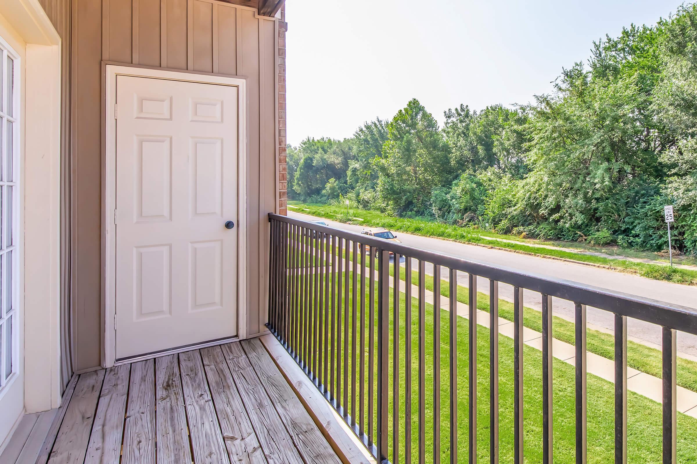 A wooden balcony with a black railing overlooking a green area filled with trees. A white door leads into an interior space, and a paved sidewalk runs adjacent to a road in the distance. The scene is bright and sunny, suggesting a peaceful outdoor setting.