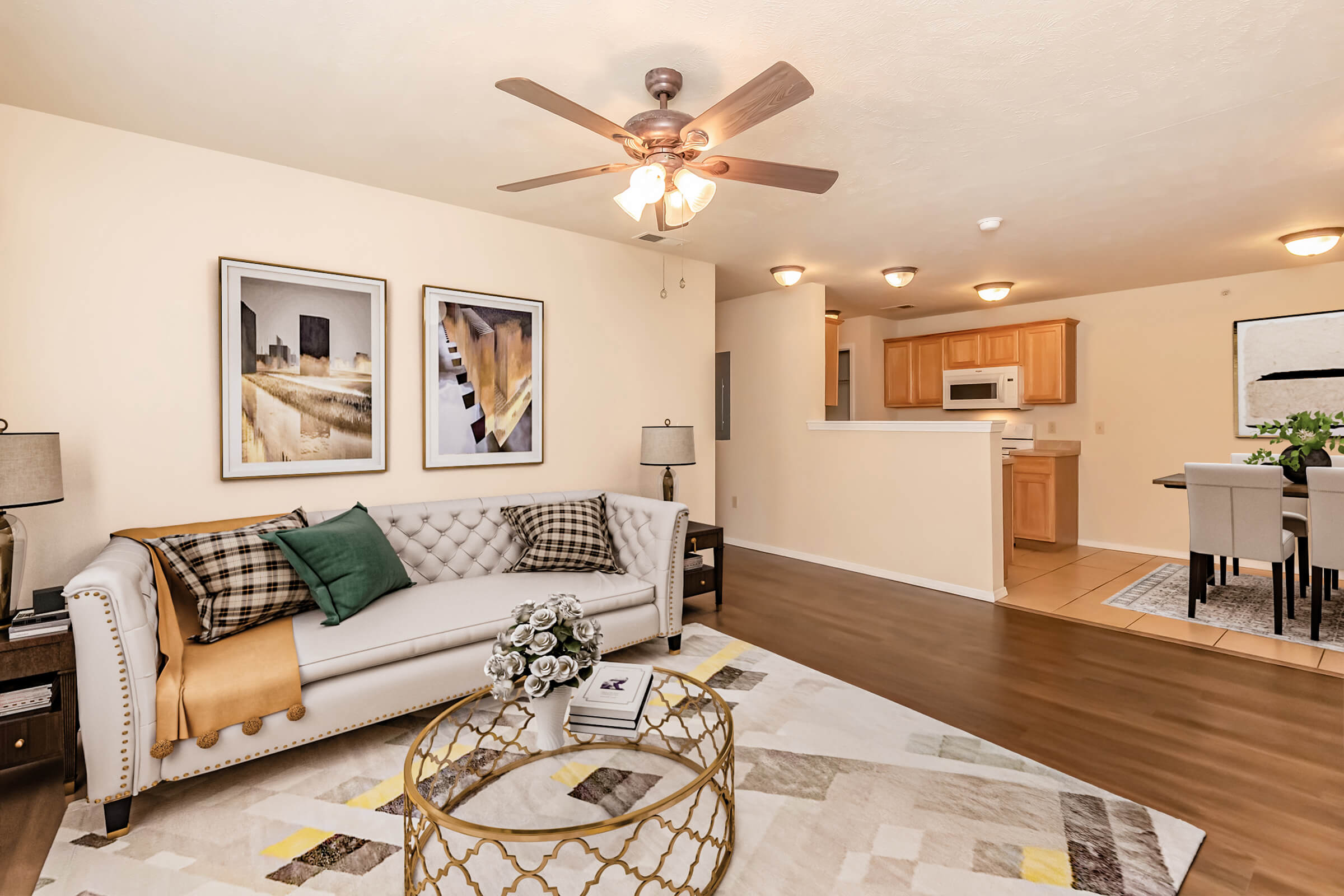 Modern living room featuring a white sofa with decorative pillows, a patterned area rug, and a coffee table. Two framed art pieces hang on the wall. A ceiling fan is visible, alongside lamps and a dining area with a kitchen in the background. The overall decor is stylish and inviting, with warm lighting.