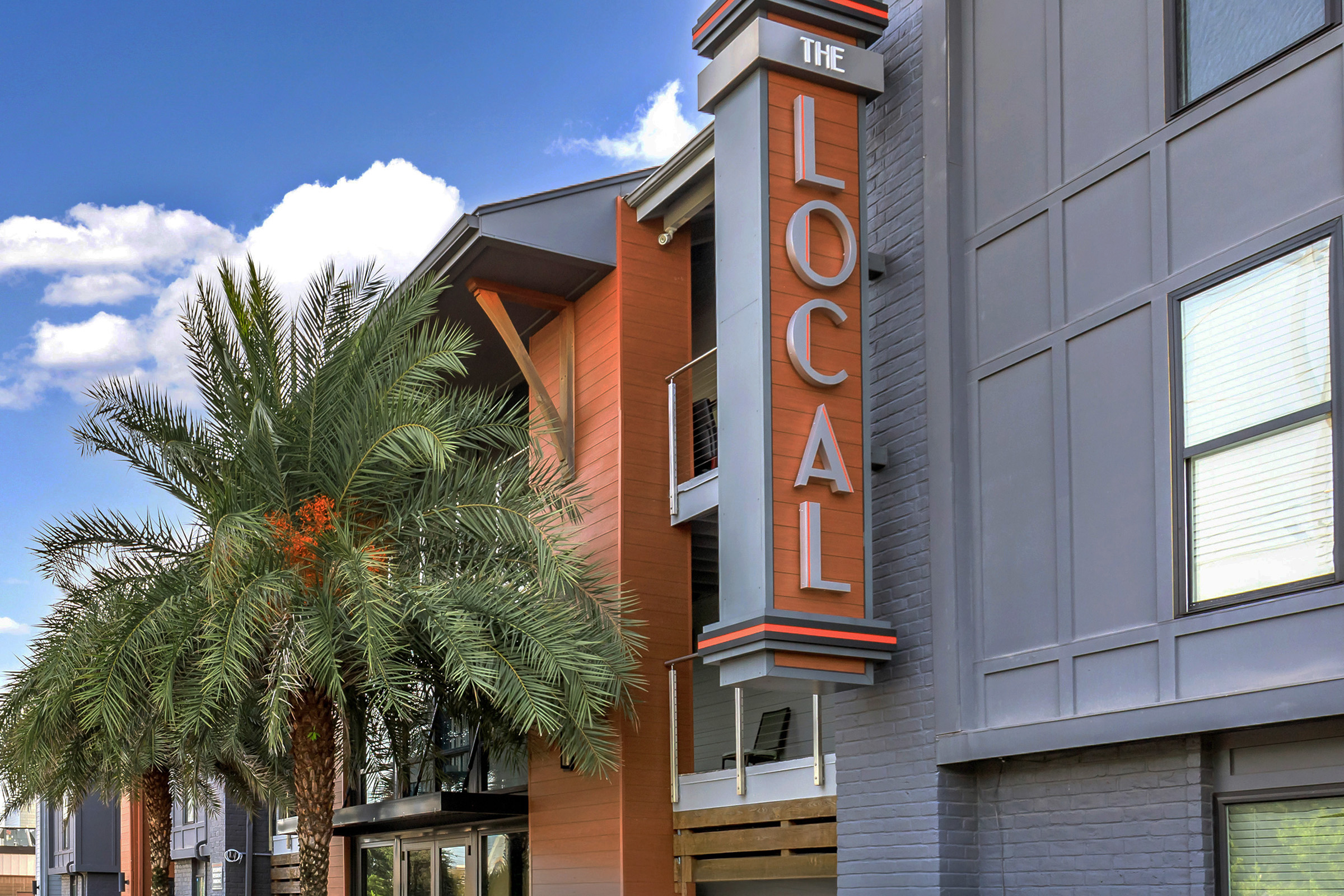 Exterior view of a building with a large sign reading "THE LOCAL." The structure features a mix of modern architecture and palm trees, set against a bright blue sky with fluffy clouds. The design includes a combination of gray and orange elements, creating a welcoming and vibrant atmosphere.