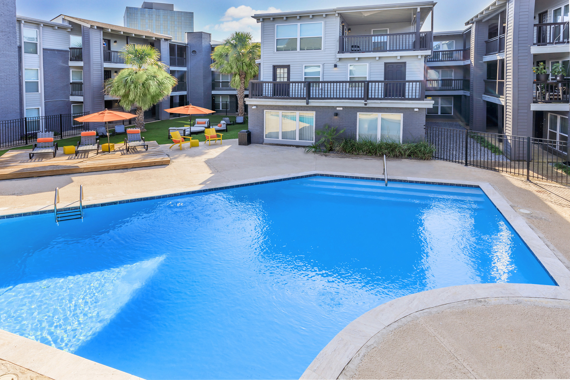A sparkling blue swimming pool surrounded by palm trees and lounge chairs. The area features orange umbrellas and a grassy space, with modern apartment buildings in the background. The scene is bright and inviting, typical of a resort-style living environment.