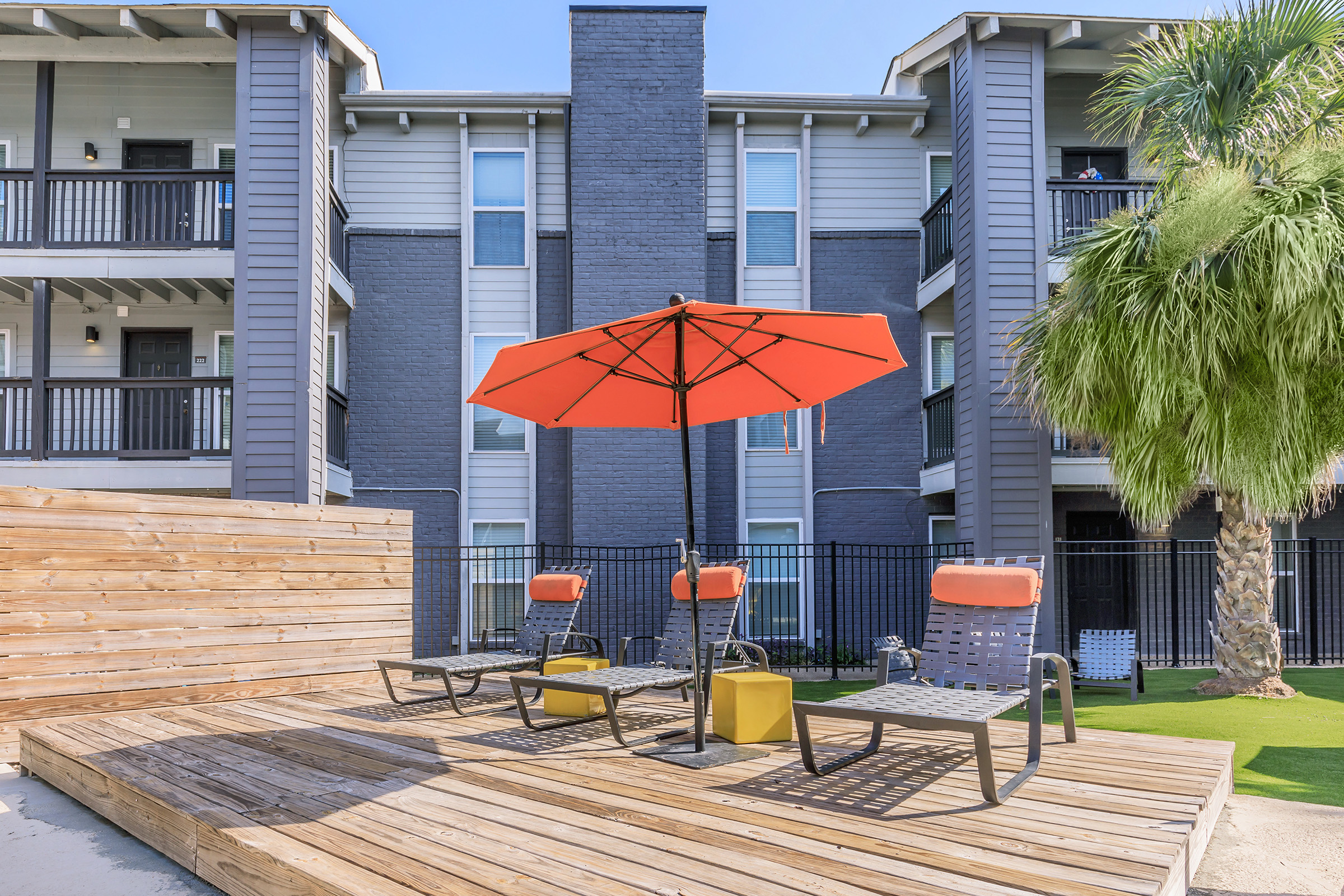 Outdoor seating area featuring two lounge chairs and a bright orange umbrella on a wooden deck. In the background, there is a multi-story apartment building with balconies and green palm trees. The setting is well-lit, suggesting a sunny day.
