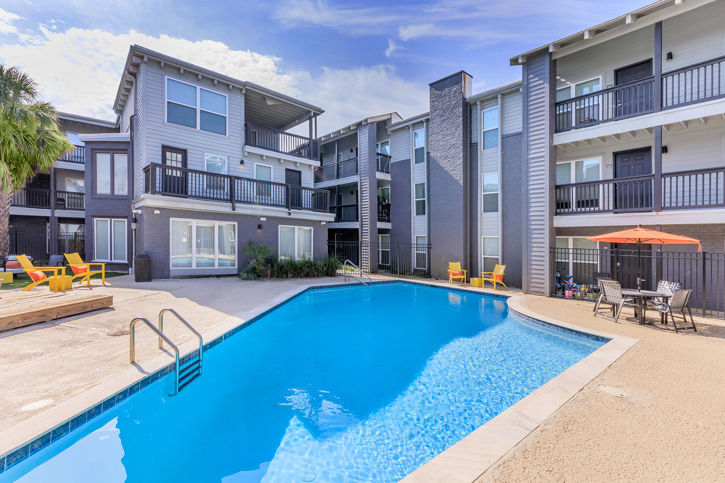Swimming pool surrounded by modern apartment buildings with balconies. Sun loungers in vibrant colors are placed near the pool, and an orange umbrella provides shade over outdoor seating. Lush greenery adds a touch of nature to the urban setting under a partly cloudy sky.