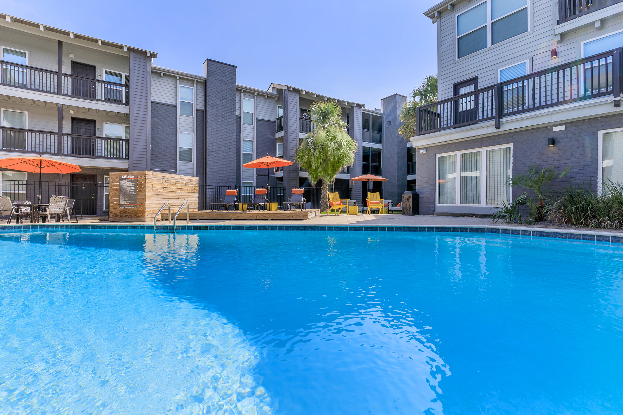 A clear blue swimming pool surrounded by lounge chairs and orange umbrellas, with modern apartment buildings in the background. Lush greenery adds to the inviting atmosphere, making it a perfect spot for relaxation and leisure. Bright, sunny weather enhances the scene.