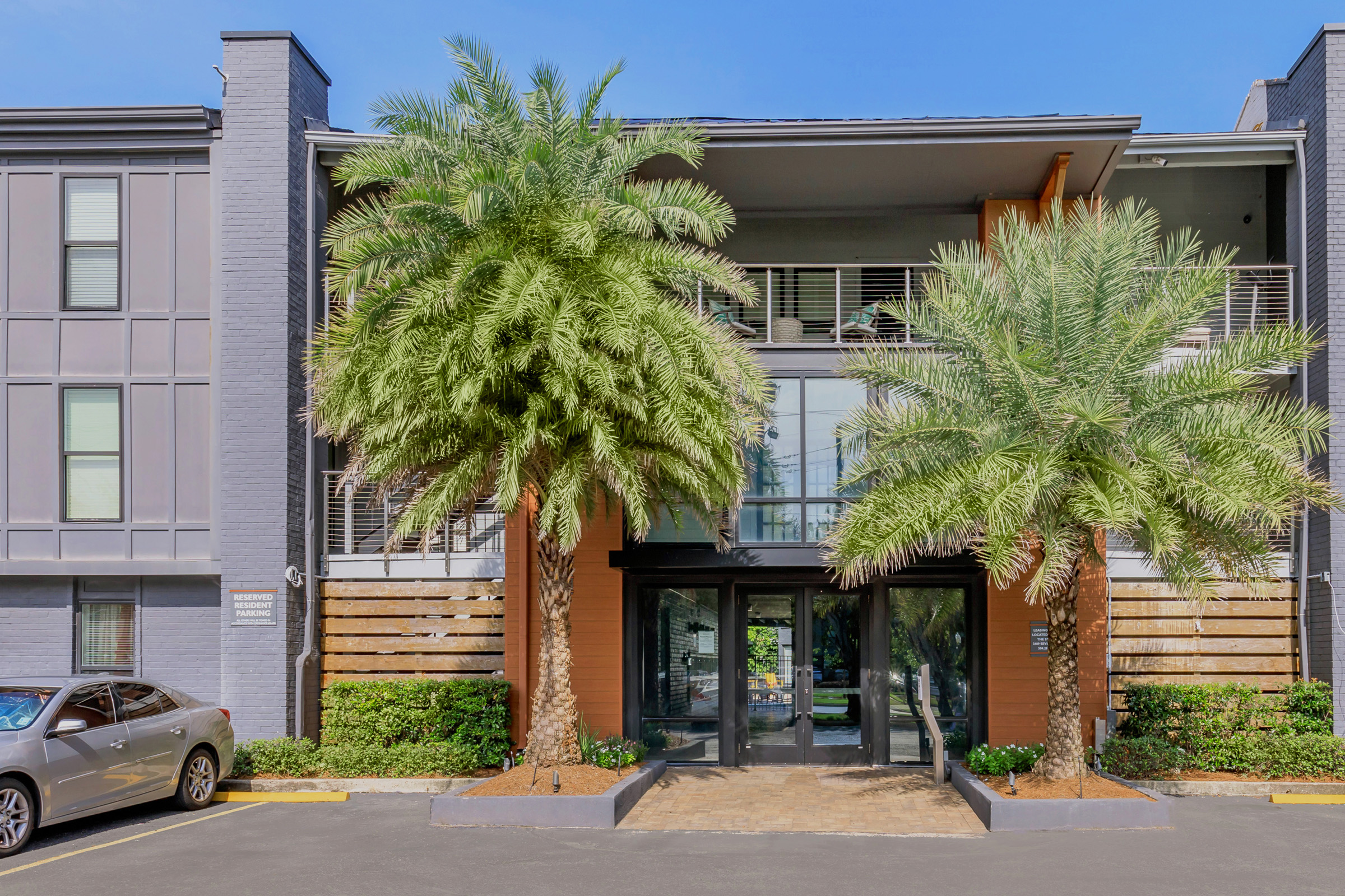 Modern apartment building entrance surrounded by palm trees, with a glass door leading inside. The exterior features a mix of gray and wooden accents, alongside neatly trimmed landscaping. A parked car is visible in the foreground, with a clear blue sky overhead.