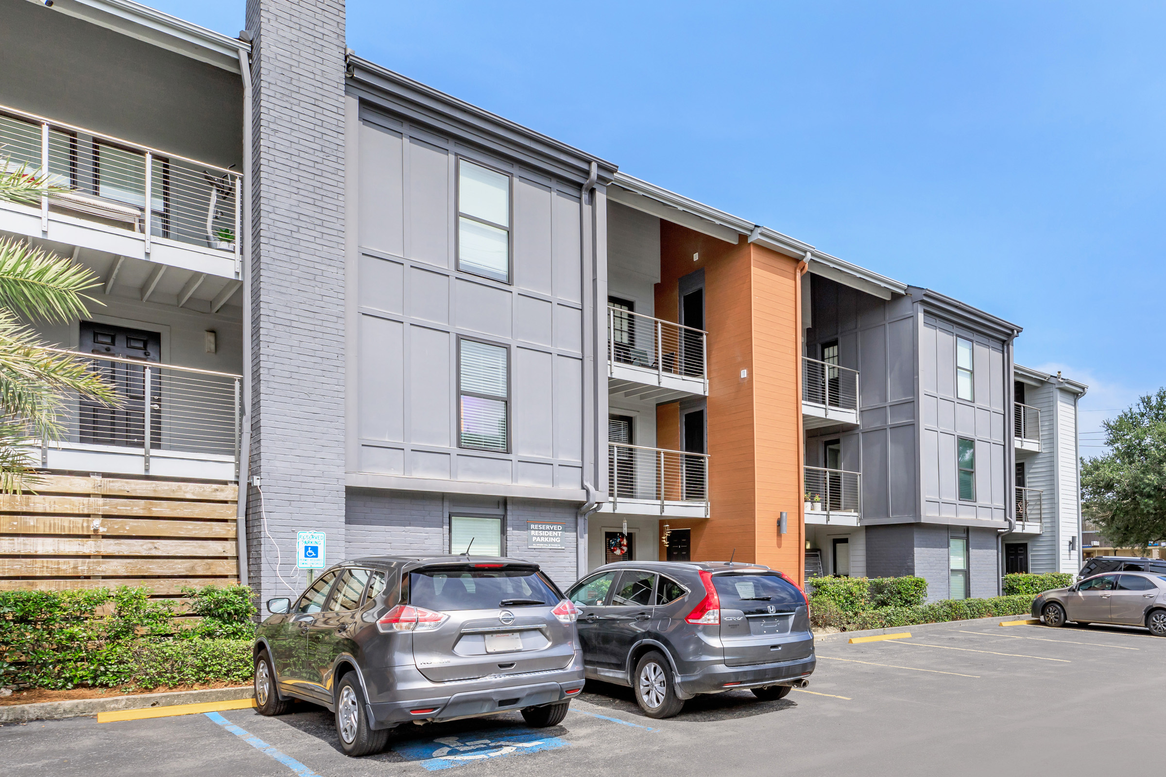 Modern apartment building with a mix of gray and orange exteriors. Two parked cars in the foreground, one gray and one dark blue. Lush greenery surrounds the area, and a clear blue sky is above. The building features balconies and clean lines, indicating a contemporary architectural style.