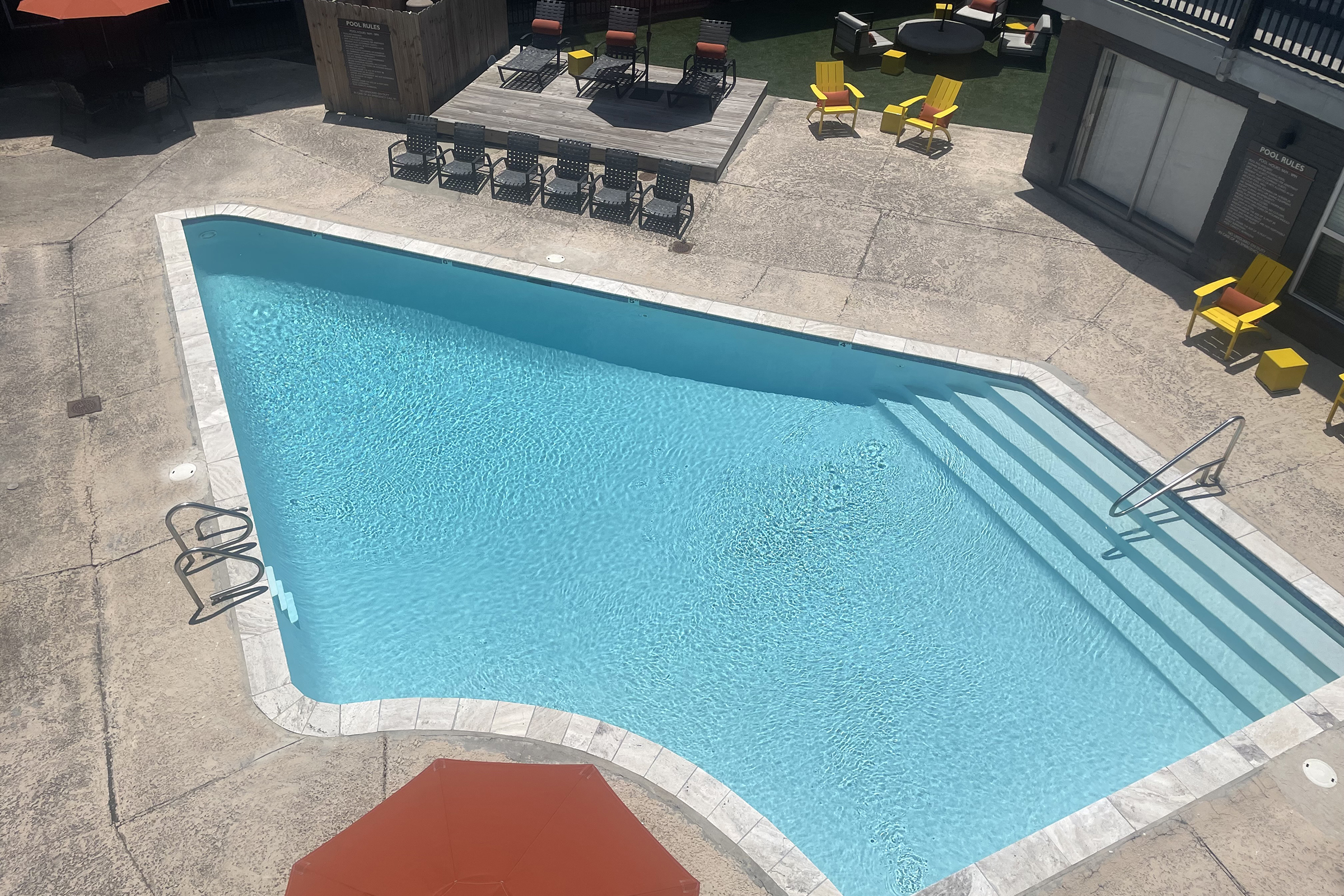 A clear, blue swimming pool surrounded by a concrete deck. There are metal ladders on one side, and several lounge chairs and bright yellow chairs are arranged around the pool area. An umbrella provides shade in one corner, and the setting appears sunny and inviting, likely in a residential or vacation complex.